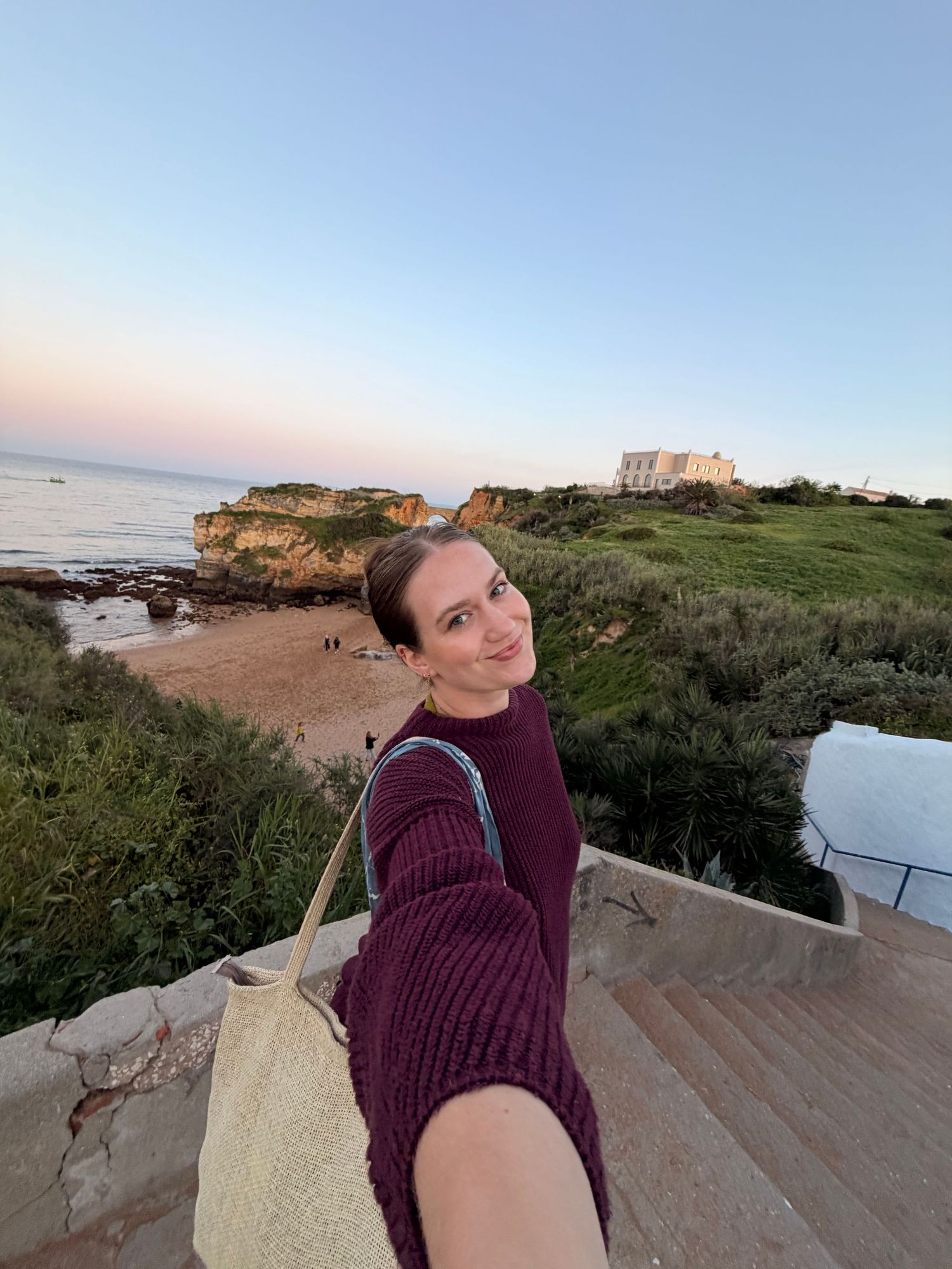 Woman in burgundy sweater takes seaside selfie at sunset with rocky coastal cliffs in background.