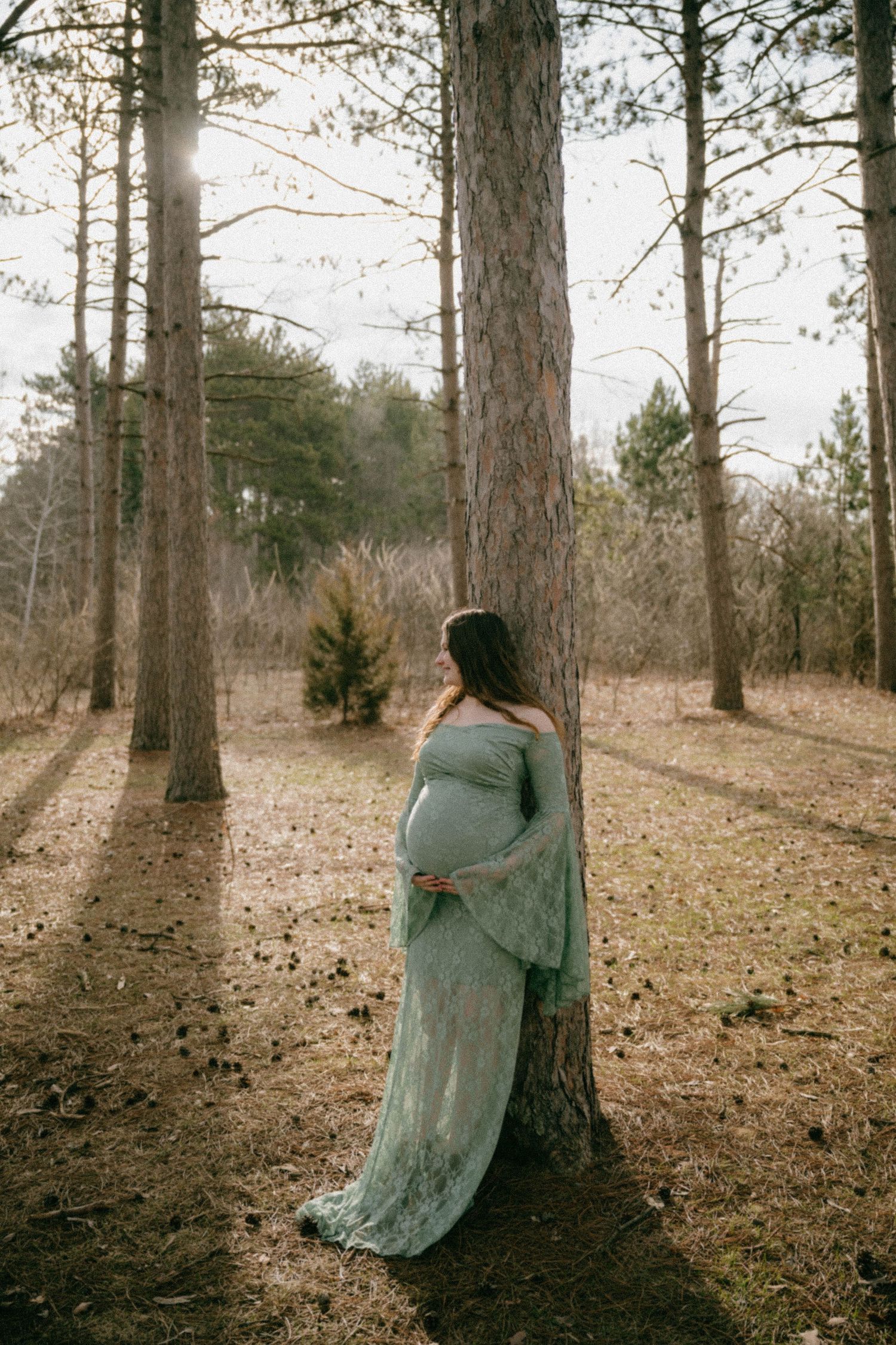 Pregnant woman in flowing teal lace gown leaning against pine tree in forest.