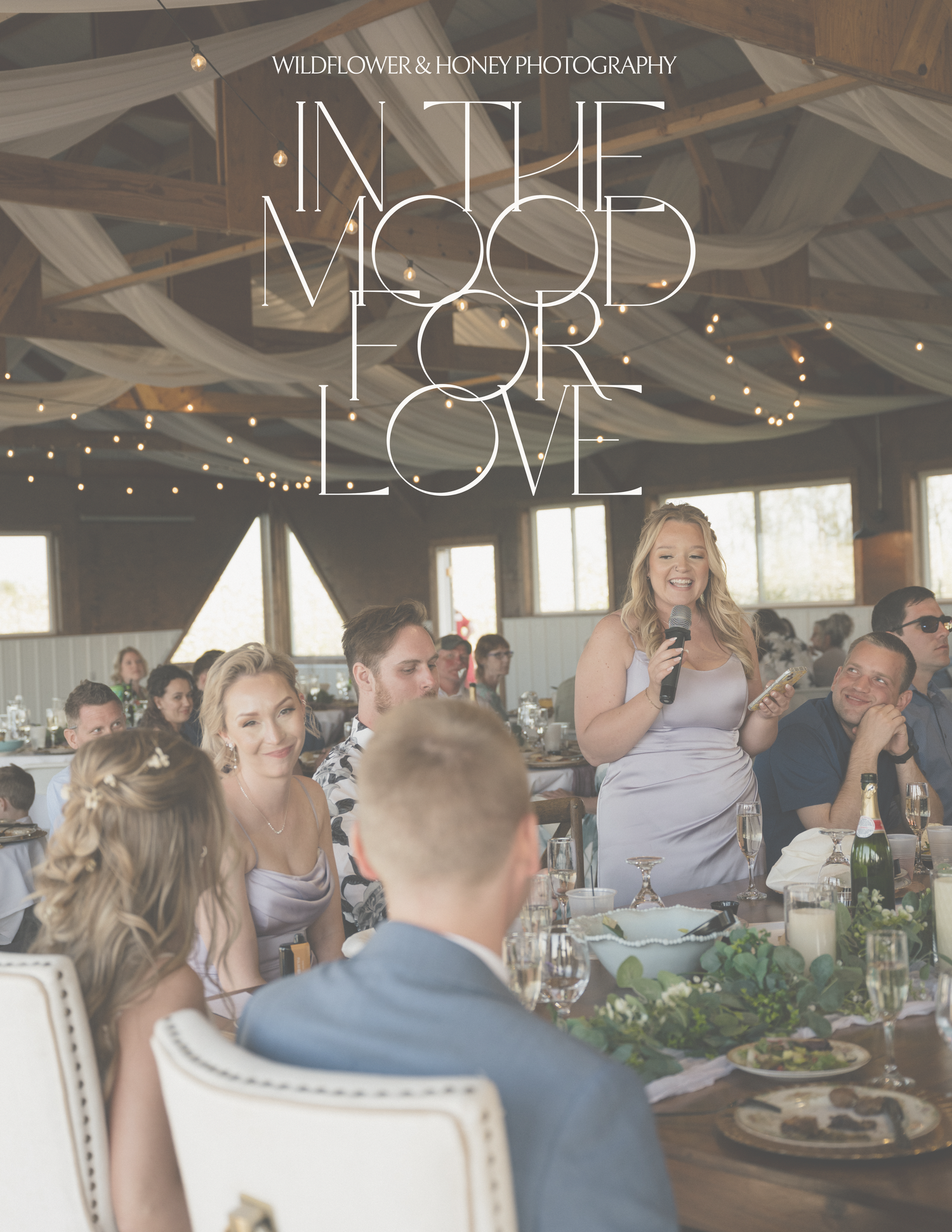 Bride laughing during wedding reception speech with guests seated at decorated tables in rustic venue.