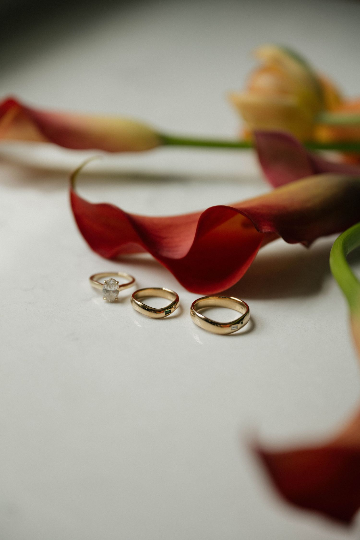 Three gold wedding rings and a diamond engagement ring arranged beside red calla lily flowers on a white surface.