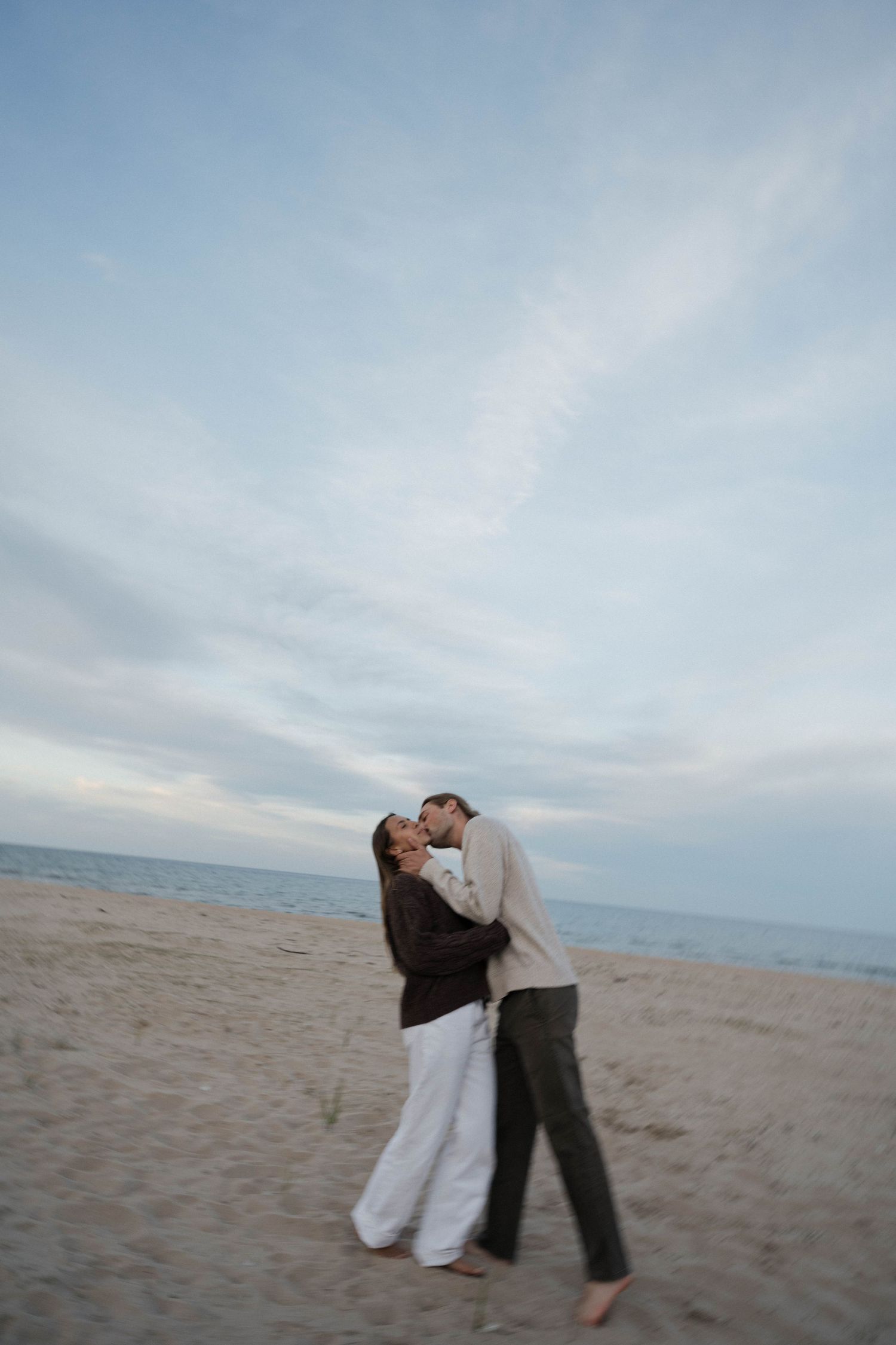 Couple sharing a romantic kiss on a sandy beach at dusk with calm ocean and cloudy sky in the background.