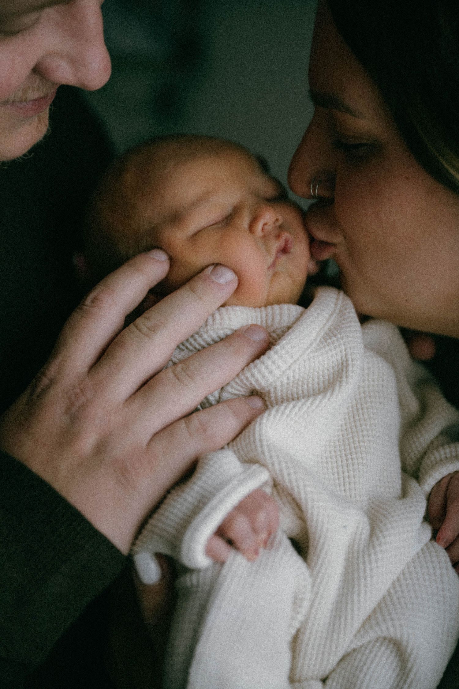 New parents tenderly holding and kissing their sleeping newborn baby wrapped in white.