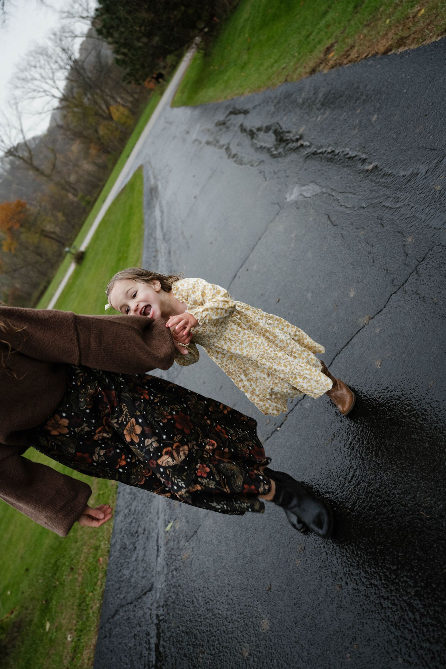 A laughing toddler in a floral dress holds her mothers hand while walking on a wet road.