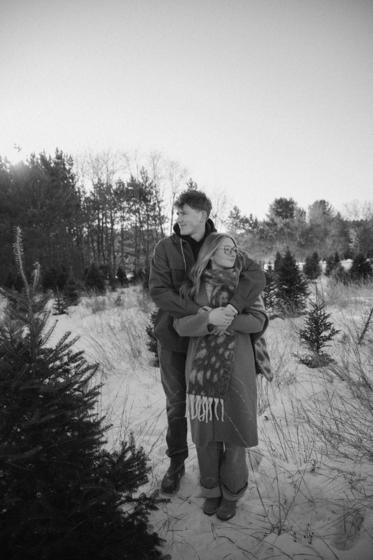 A couple embraces in a black and white photo at a snowy Christmas tree farm surrounded by pine trees.