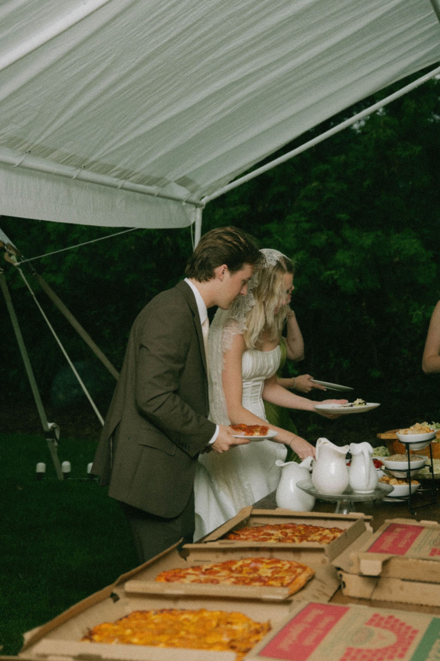 Bride and groom serving themselves pizza from open boxes at an outdoor wedding reception under a white tent.