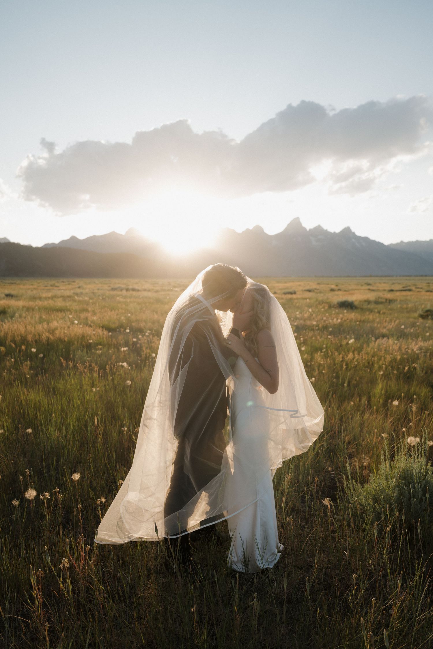 Bride and groom kissing under veil at sunset in mountain meadow with Teton peaks behind.