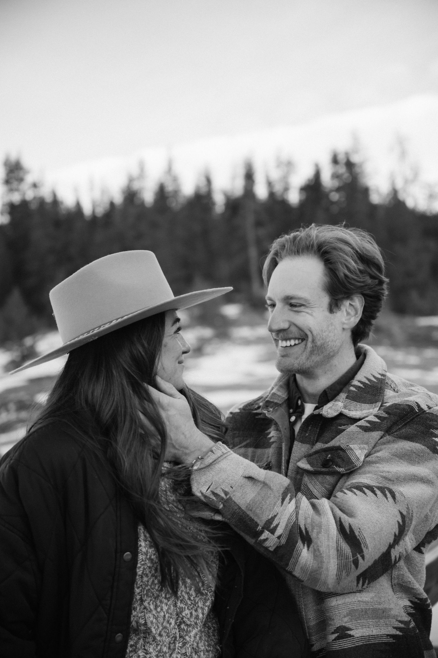 Black and white photo of smiling man tenderly touching womans face wearing wide brim hat outdoors.