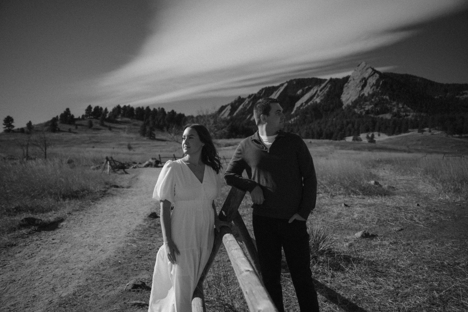 Black and white photo of engaged couple leaning on fence with dramatic Flatirons Boulder Colorado backdrop.