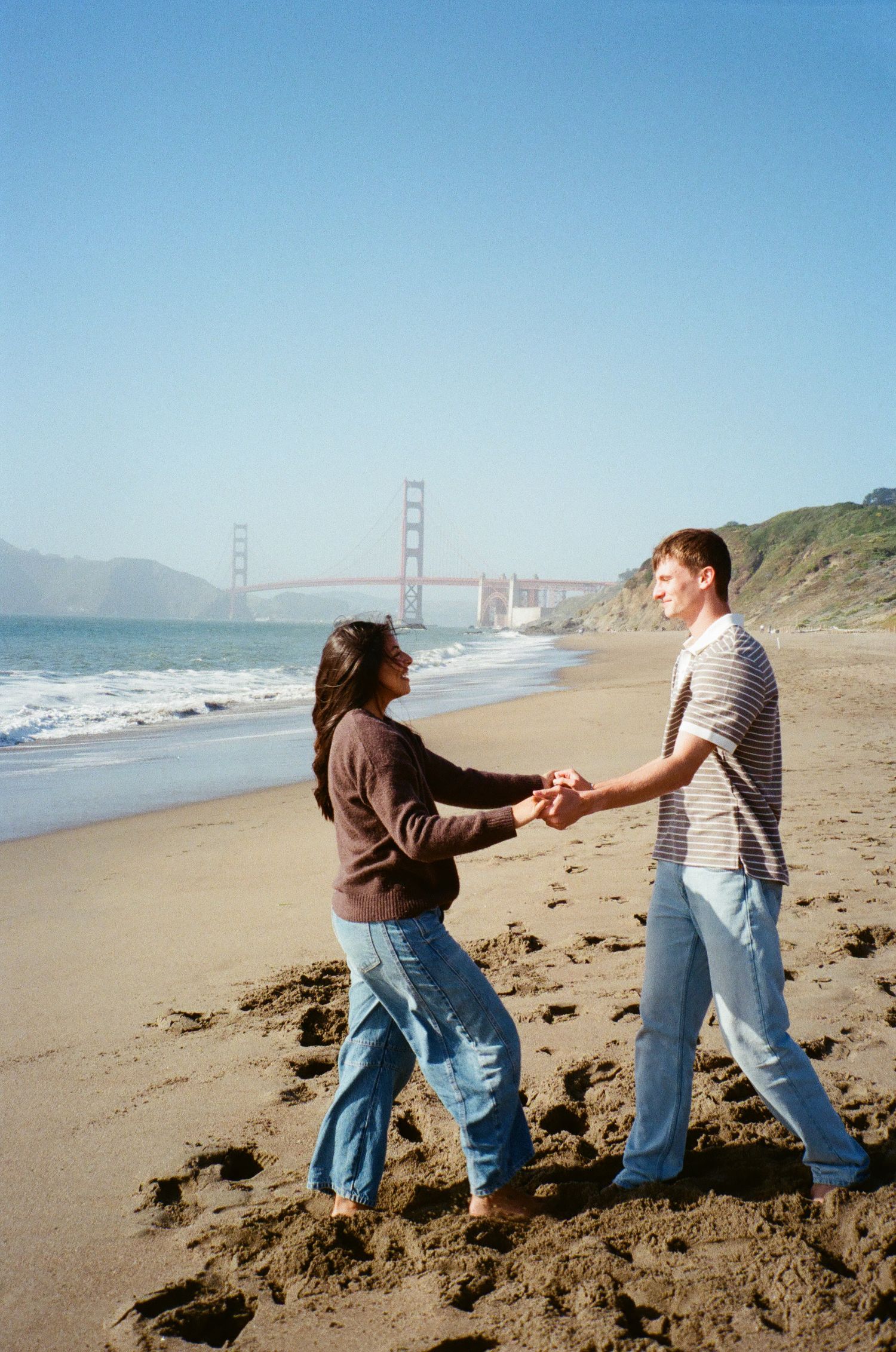 Couple holding hands and dancing on a sandy beach with the Golden Gate Bridge visible in the background.