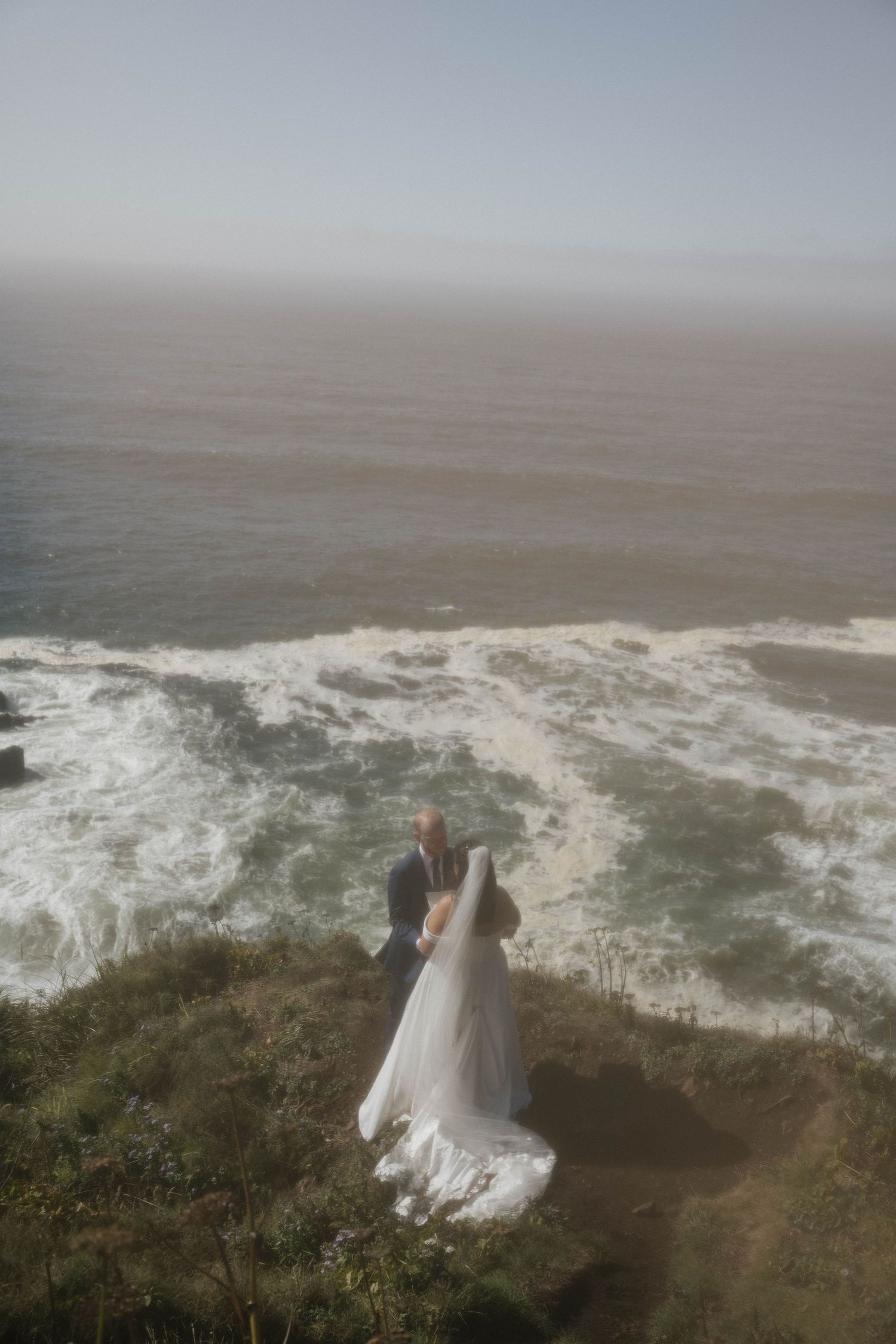 Bride in flowing white gown and groom stand on a clifftop overlooking crashing ocean waves in misty light.