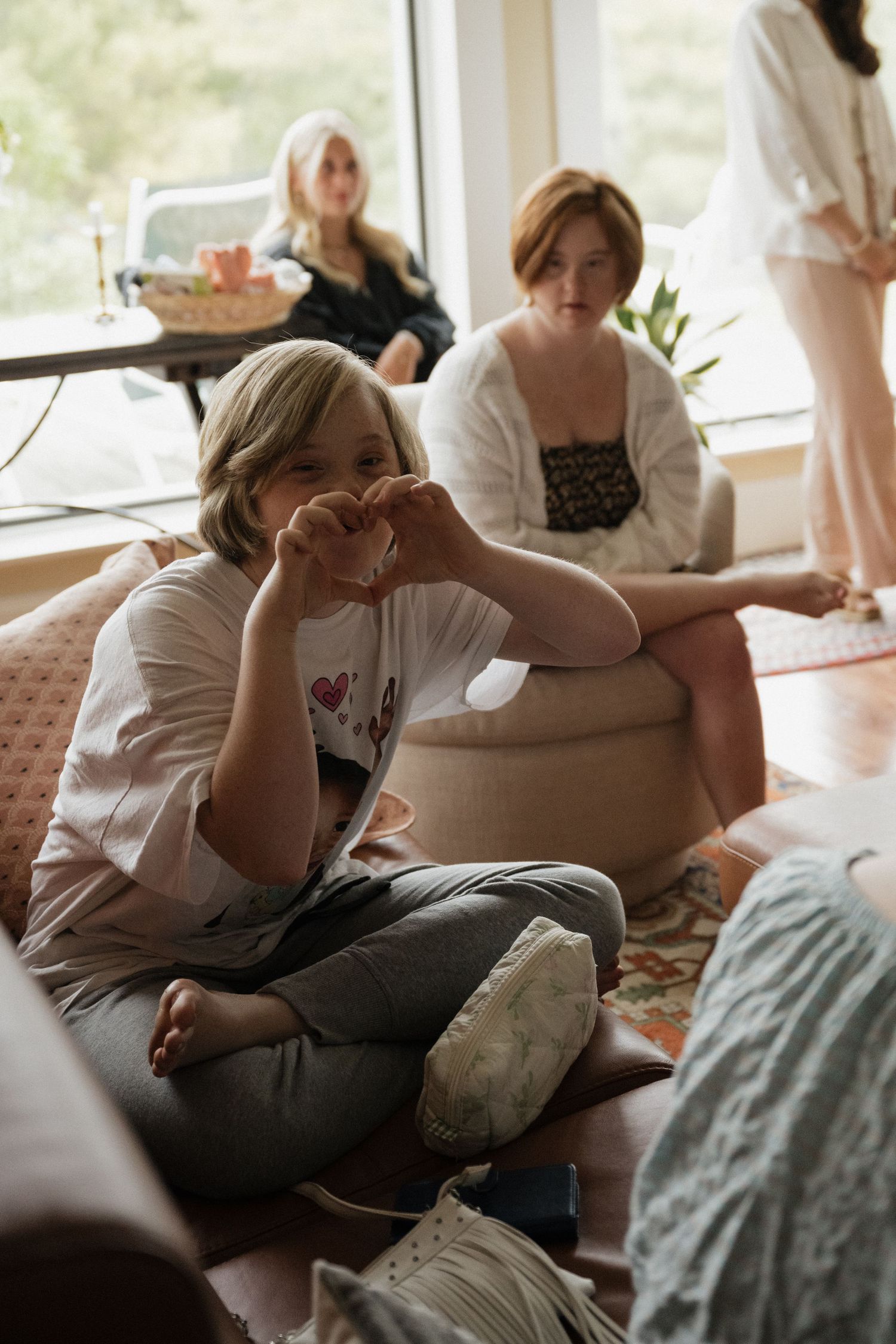 Young girl making a heart shape with her hands while sitting cross-legged on a sofa at a wedding.