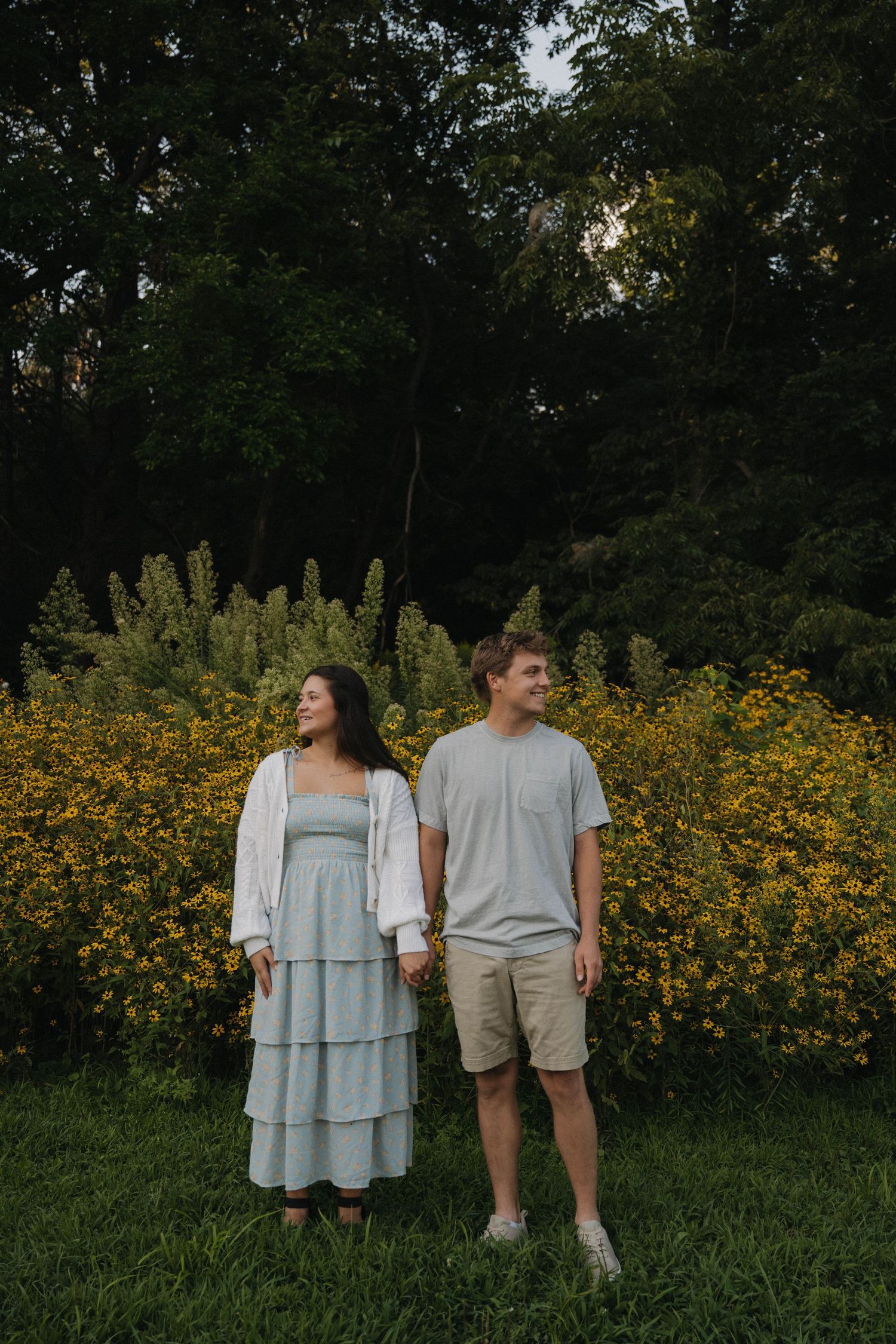 Young couple holding hands in front of yellow wildflowers and green trees at golden hour.