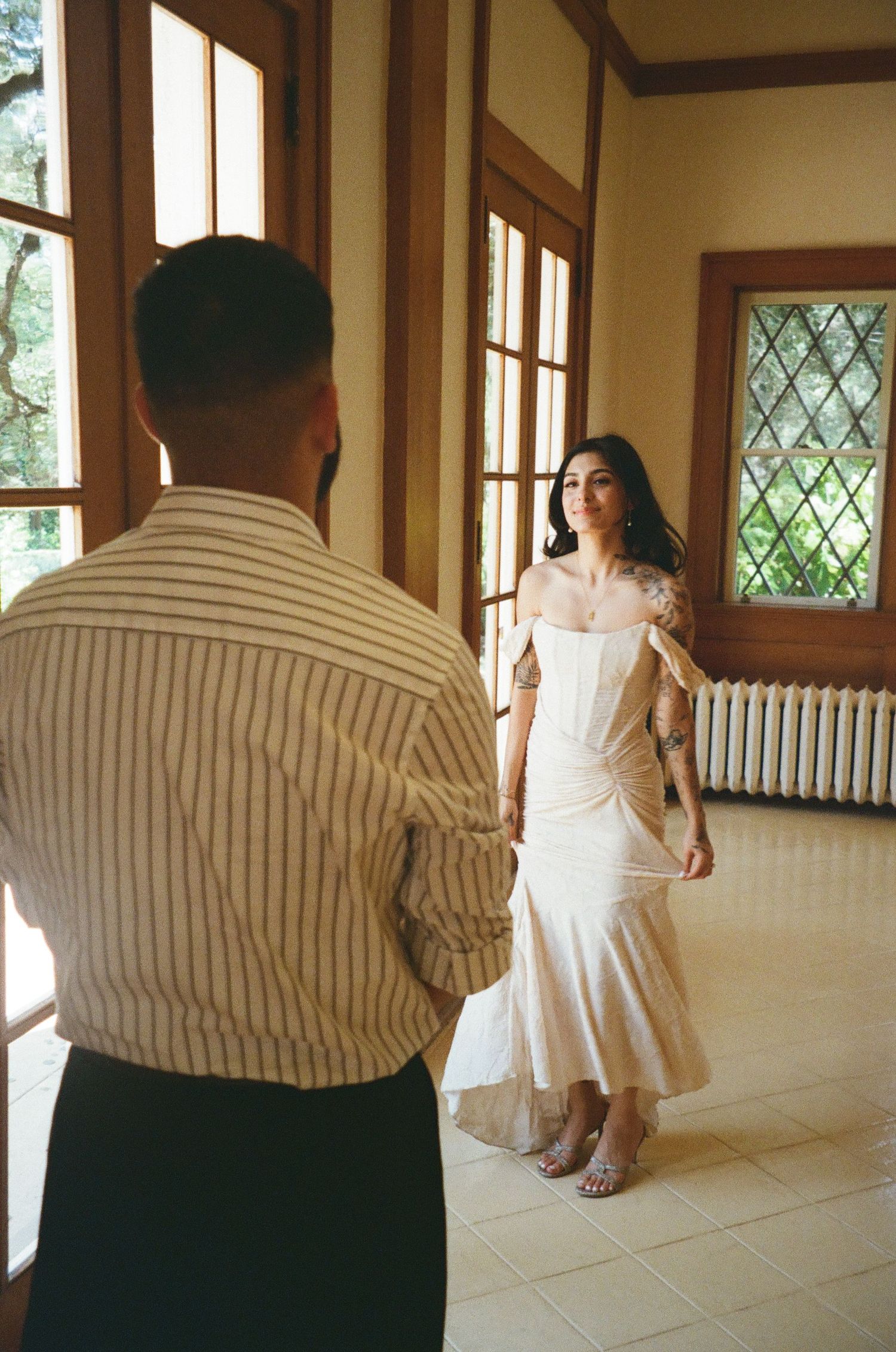 Bride in off-shoulder white gown dancing in sunlit room with groom watching during wedding.