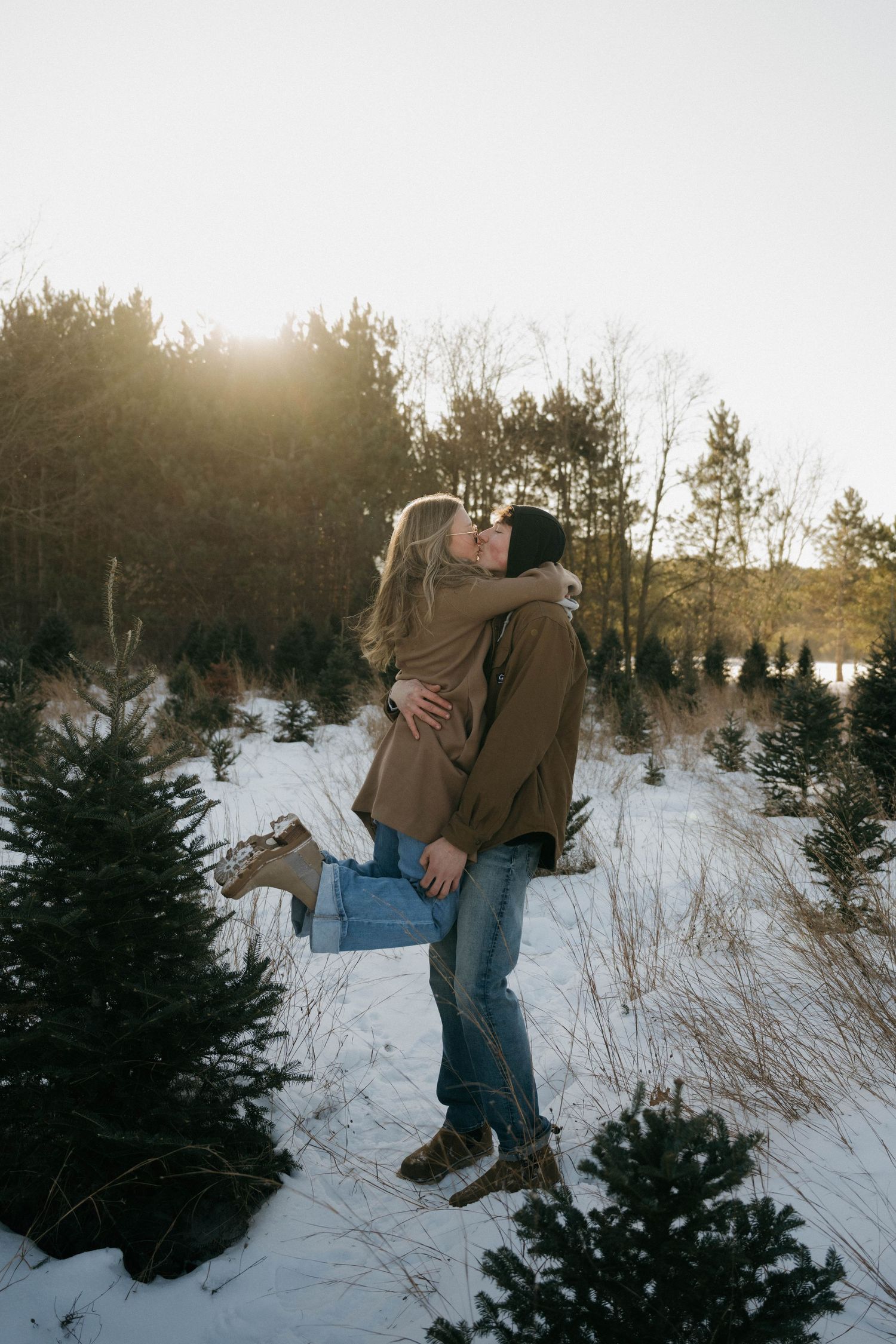 Couple sharing a romantic kiss in a snowy Christmas tree farm at golden hour.