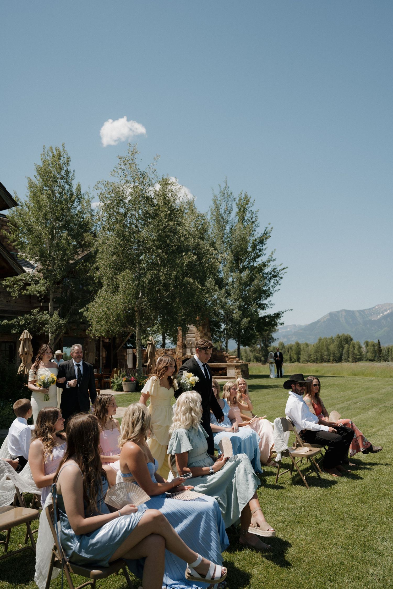 Outdoor wedding ceremony with guests seated on chairs in a mountain meadow under clear blue skies.