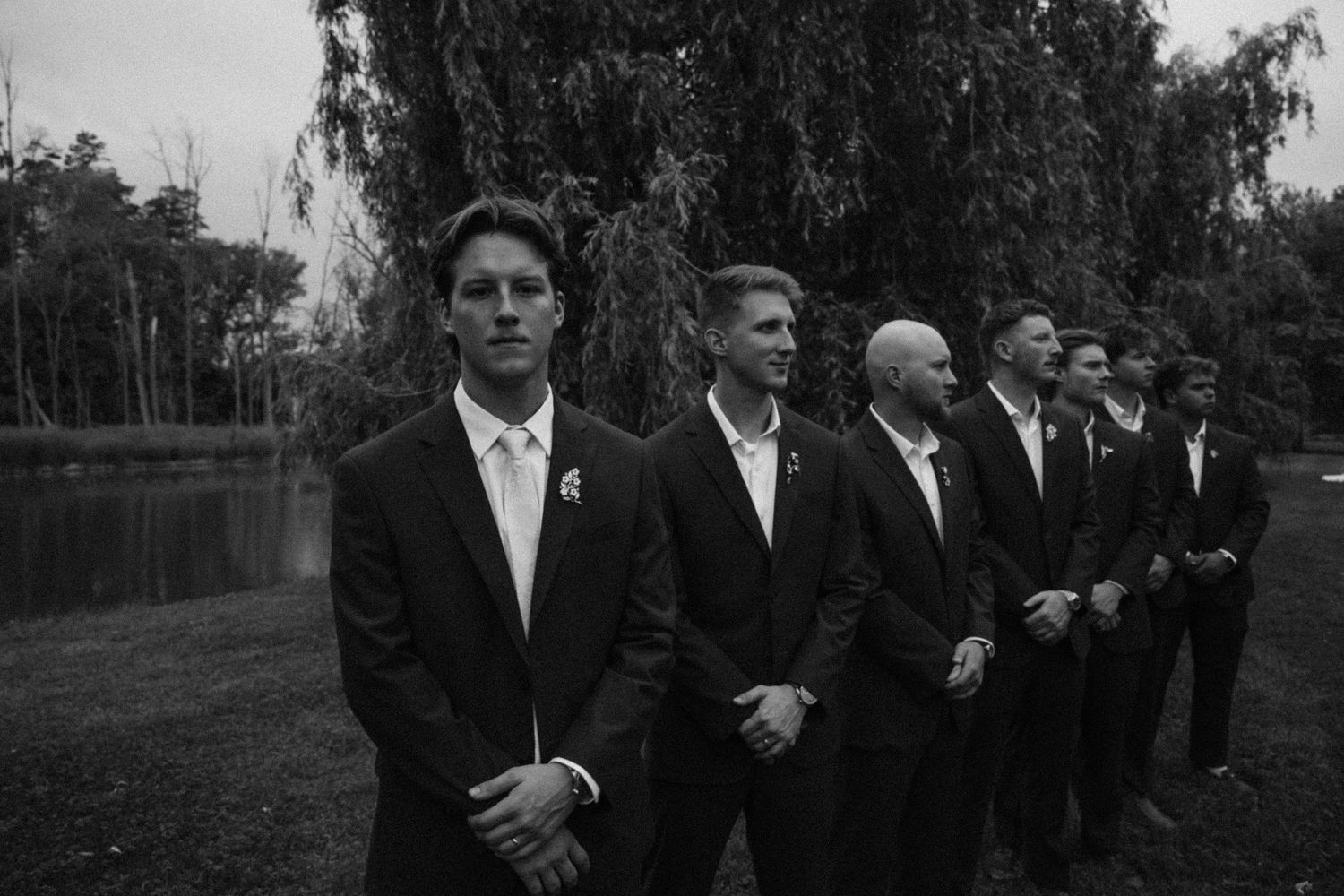 Black and white photo of groomsmen in suits standing outdoors near trees and a pond at a wedding.