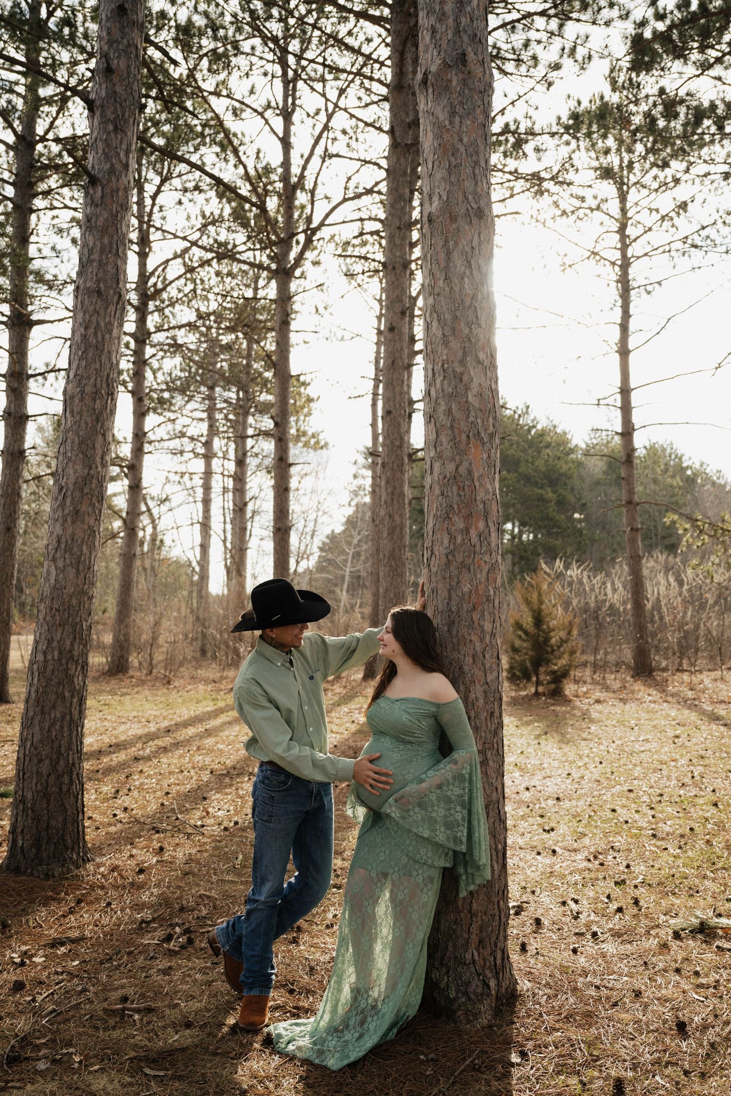 Couple in pine forest, man in cowboy hat tenderly touching pregnant woman's face.