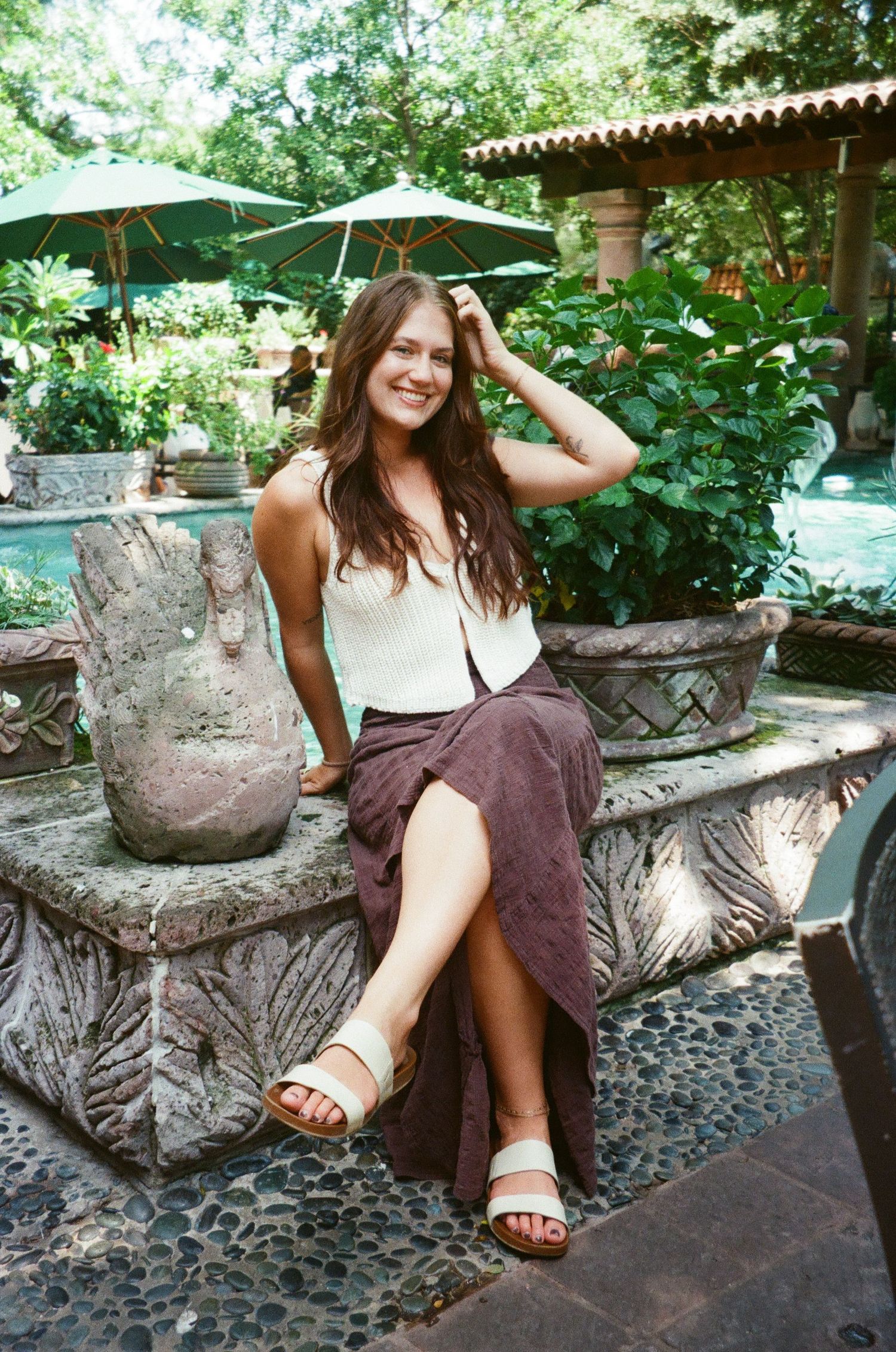 Young woman in white top and mauve skirt sitting on stone fountain in lush garden patio with pool.