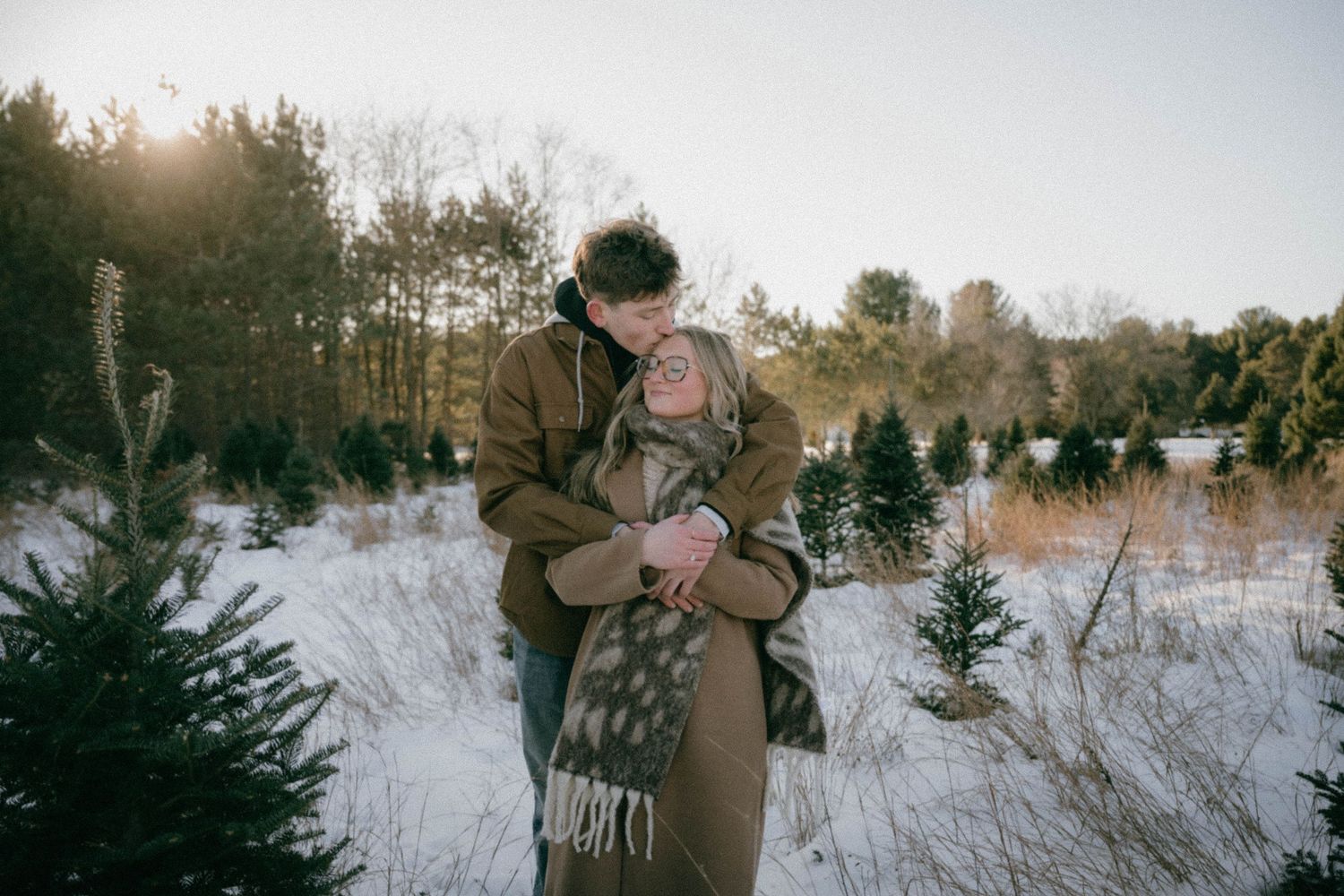Couple embraces warmly in a snowy Christmas tree farm at golden sunset.