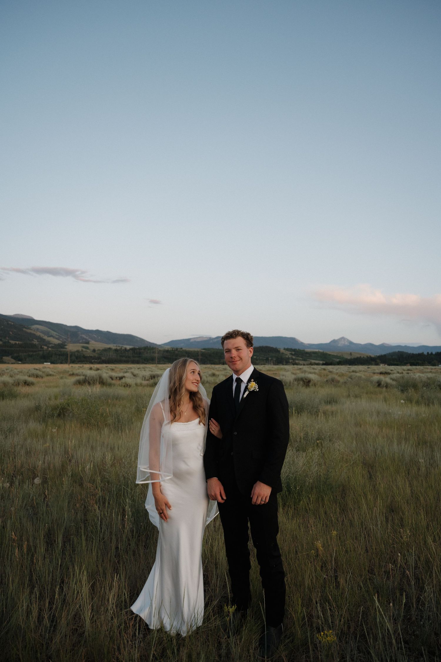 Smiling bride and groom pose in lush green meadow with mountain landscape at twilight.
