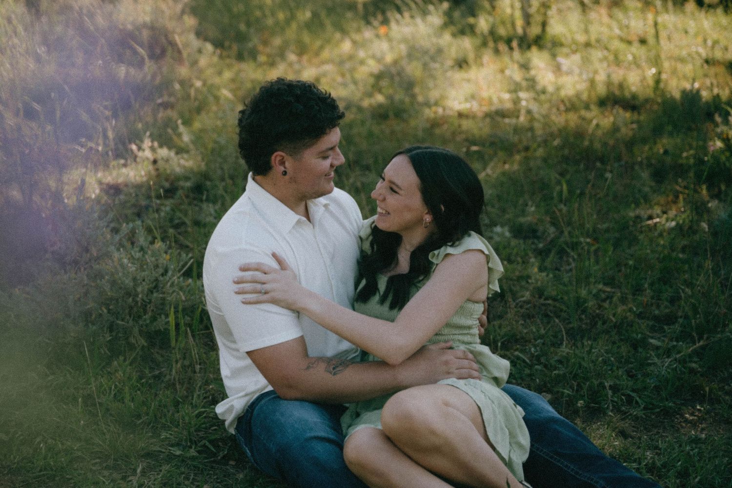 Couple sitting in sunlit meadow, smiling and embracing in warm golden outdoor light.