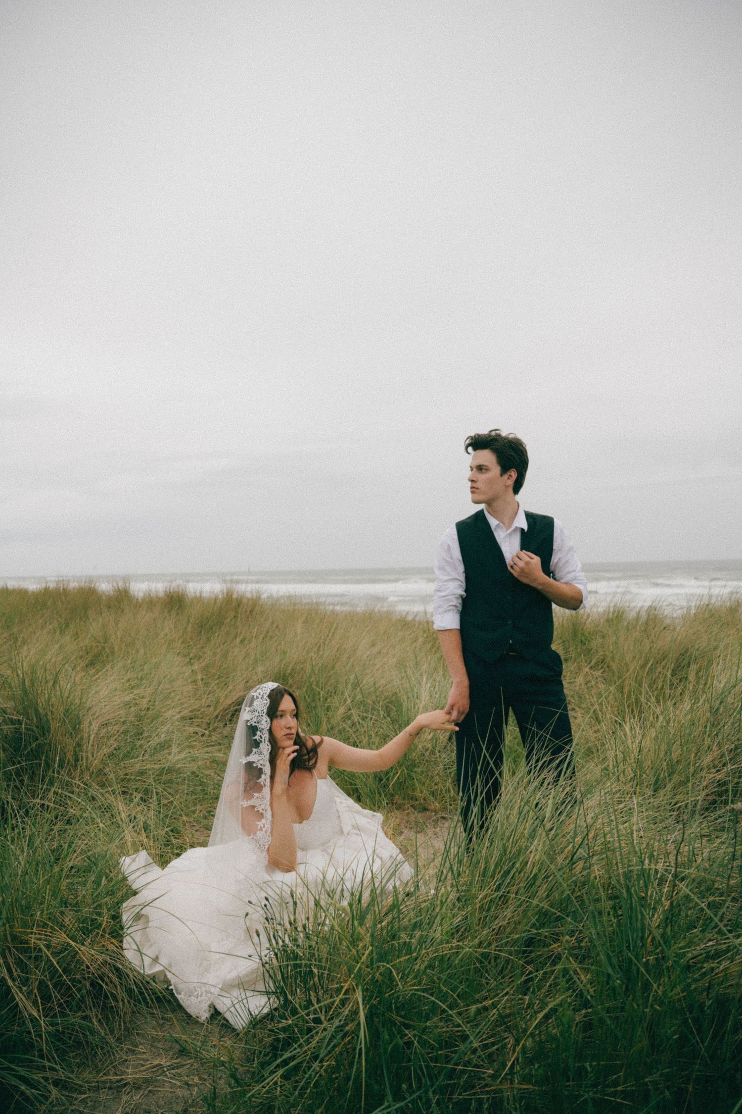 Bride in white gown and veil sits in tall beach grass holding grooms hand at coastal wedding.