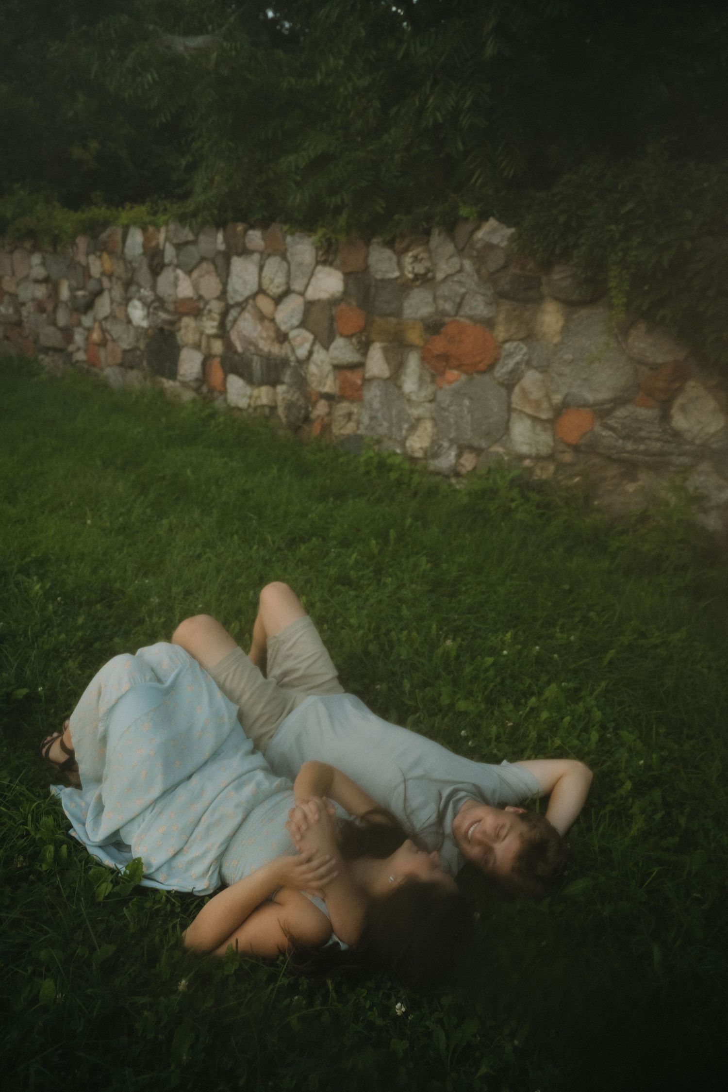 Couple relaxing on lush green grass near rustic stone wall, woman in light blue dress.