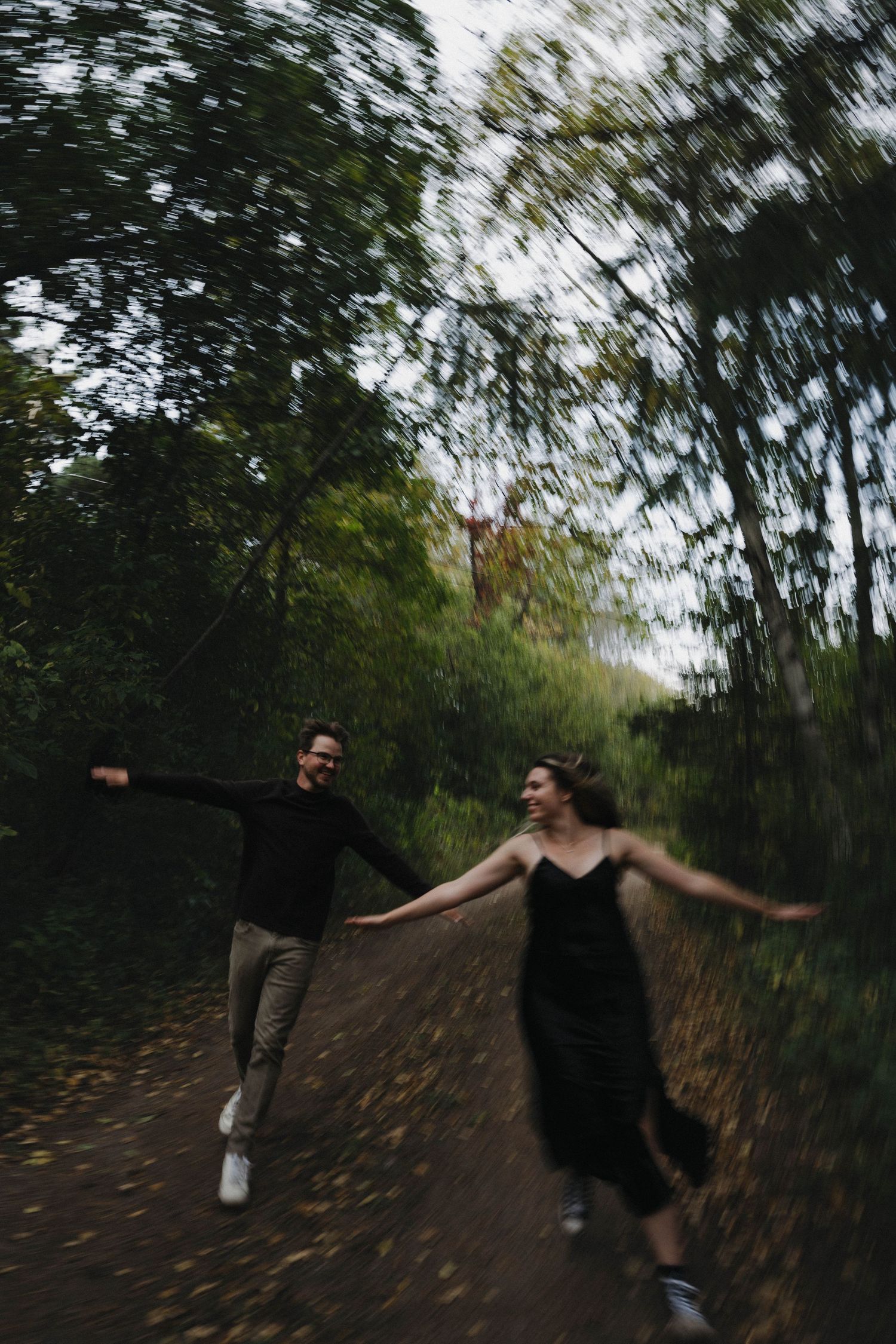 A laughing couple holding hands and running together on a leaf-covered path in a blurred forest.