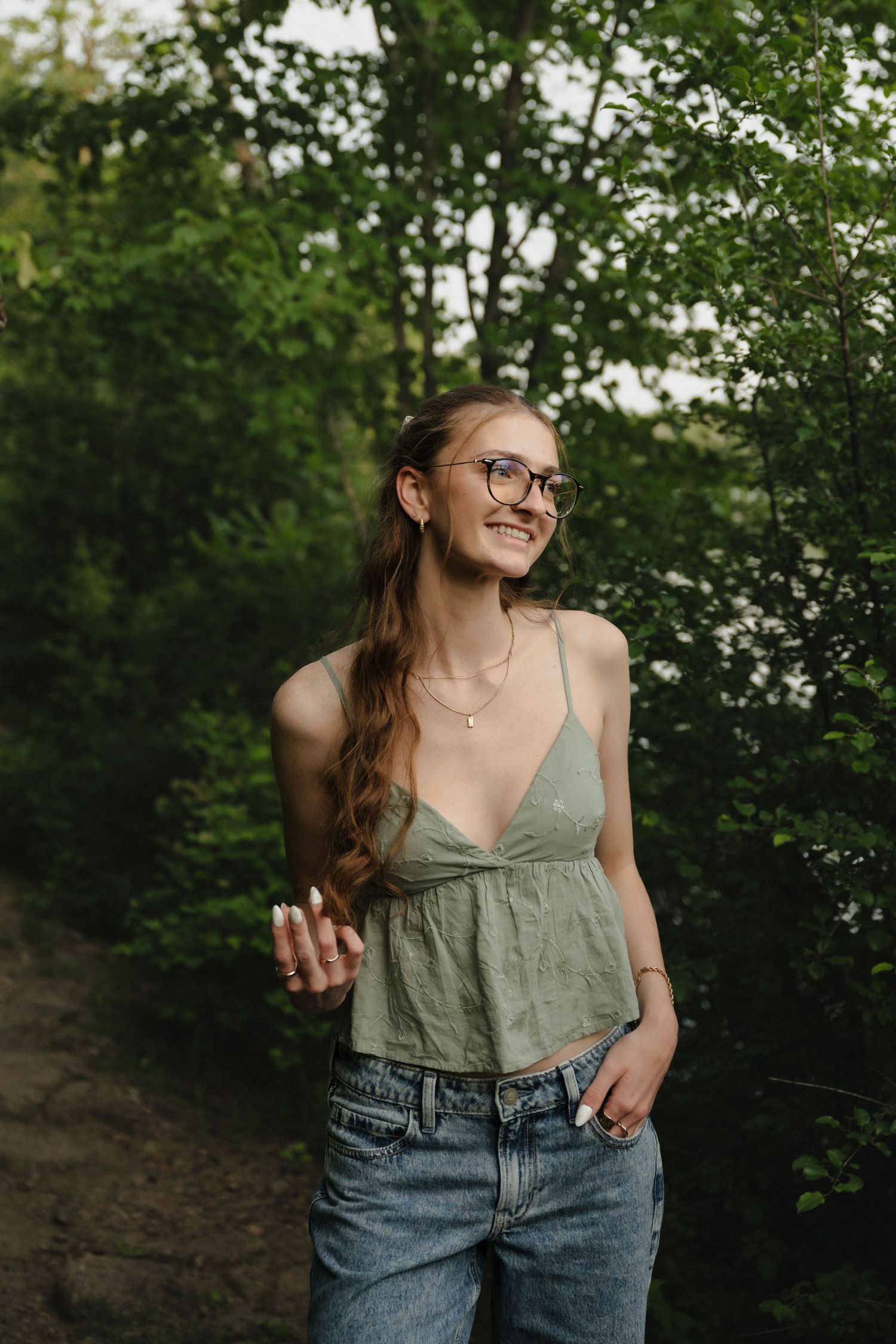 Young woman in green crop top and jeans smiles while standing on a wooded trail among green trees.