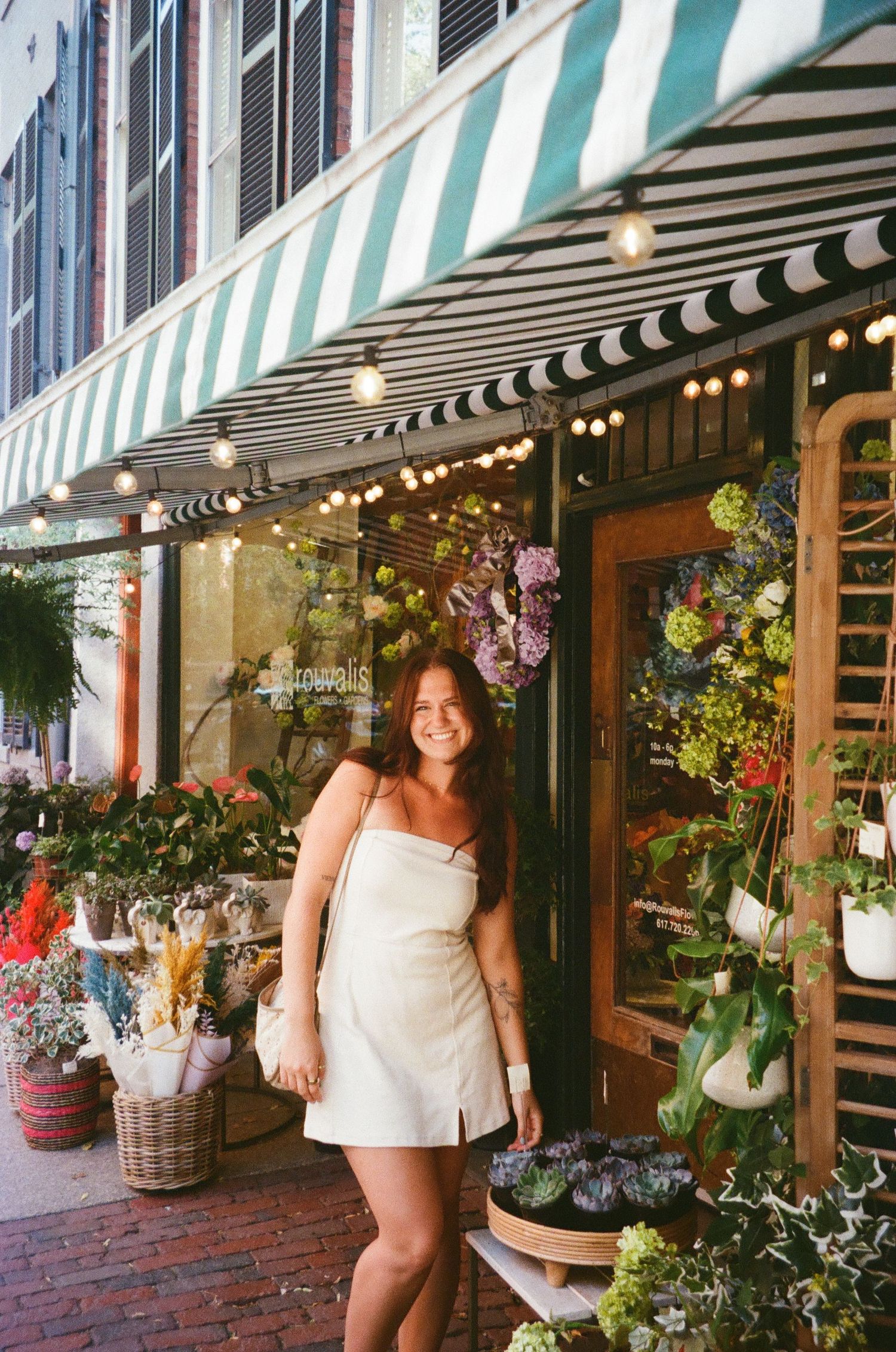 Smiling woman in white dress standing outside flower shop with striped awning and string lights.
