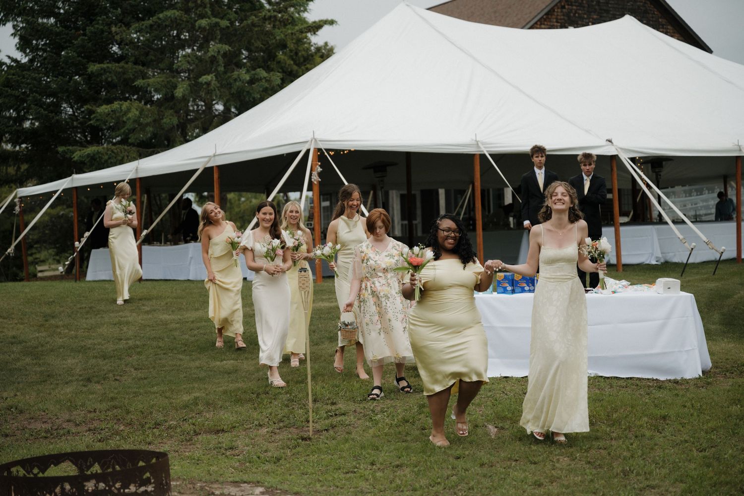 Bridesmaids in yellow dresses walk toward white reception tent outdoors.