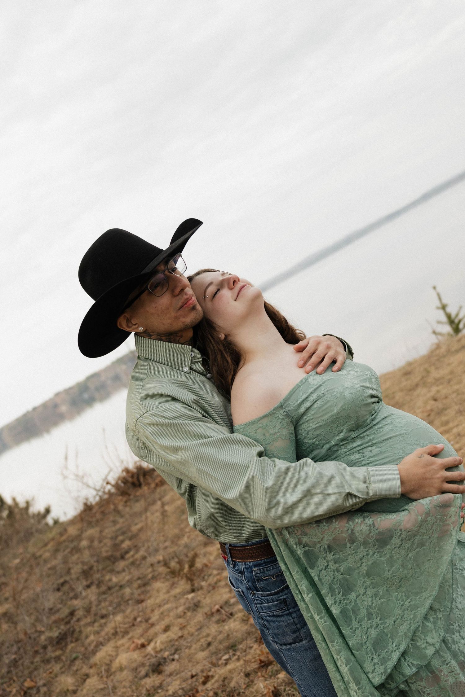 Man in black cowboy hat kissing pregnant woman's cheek in outdoor field setting.