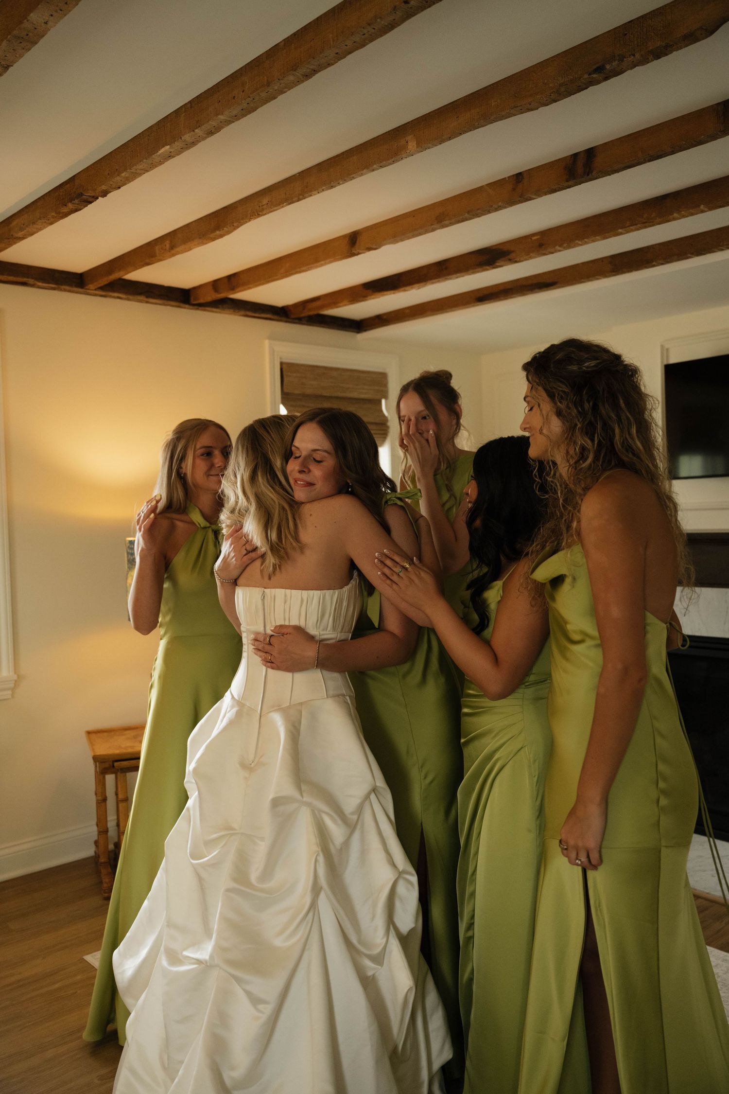 Bride in white strapless gown hugged by bridesmaids in olive green dresses inside a rustic room with wooden beam ceiling.