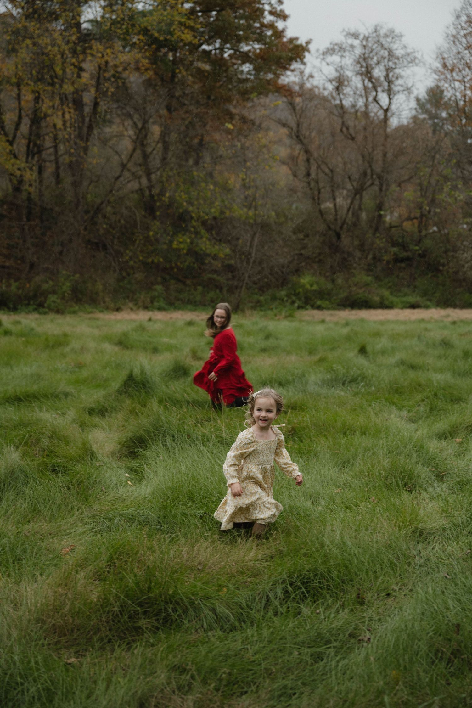 Two young girls in colorful dresses running and laughing through a lush green meadow during autumn.