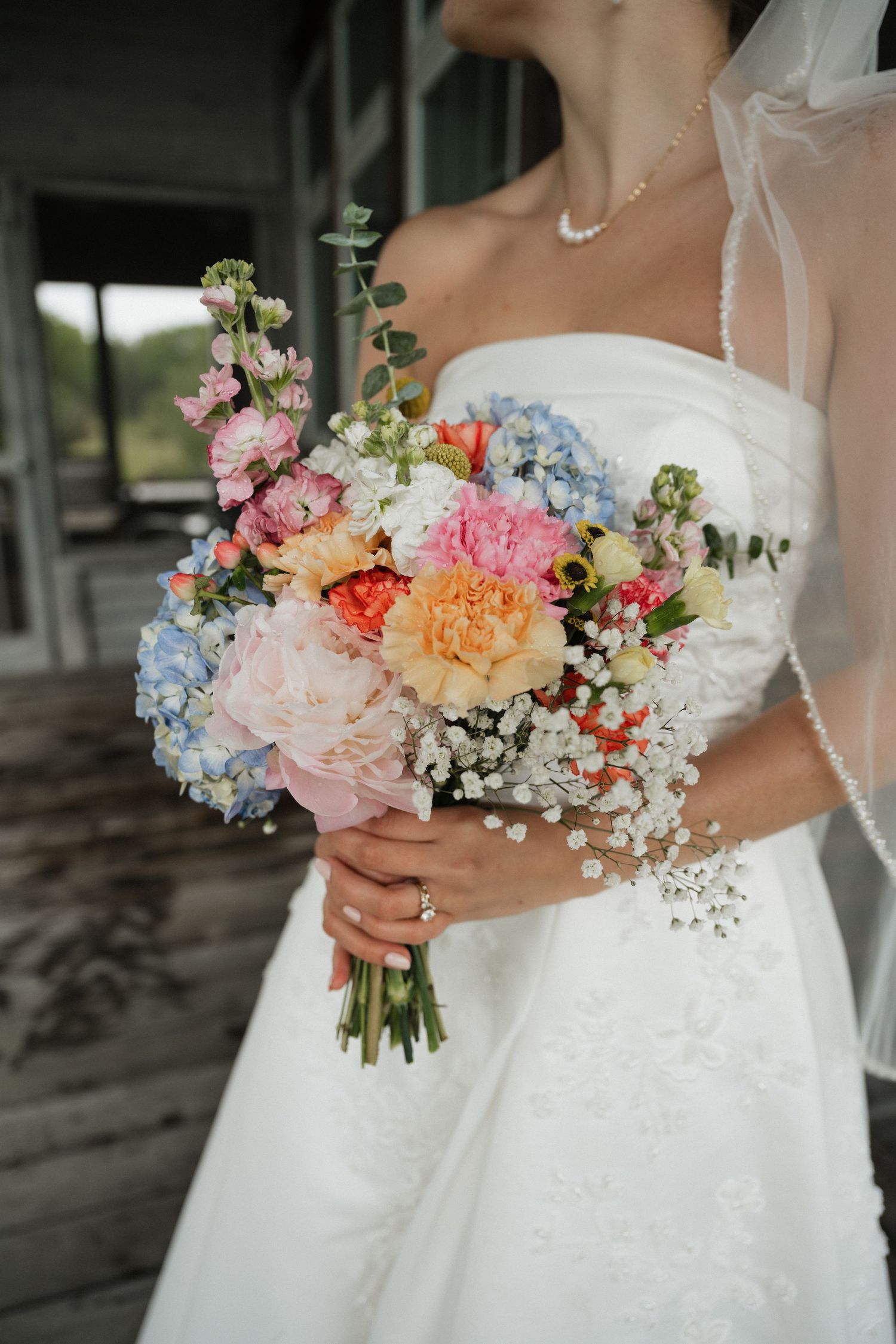 A bride in a white strapless gown holds a colorful bridal bouquet with peonies, hydrangeas, and baby's breath.