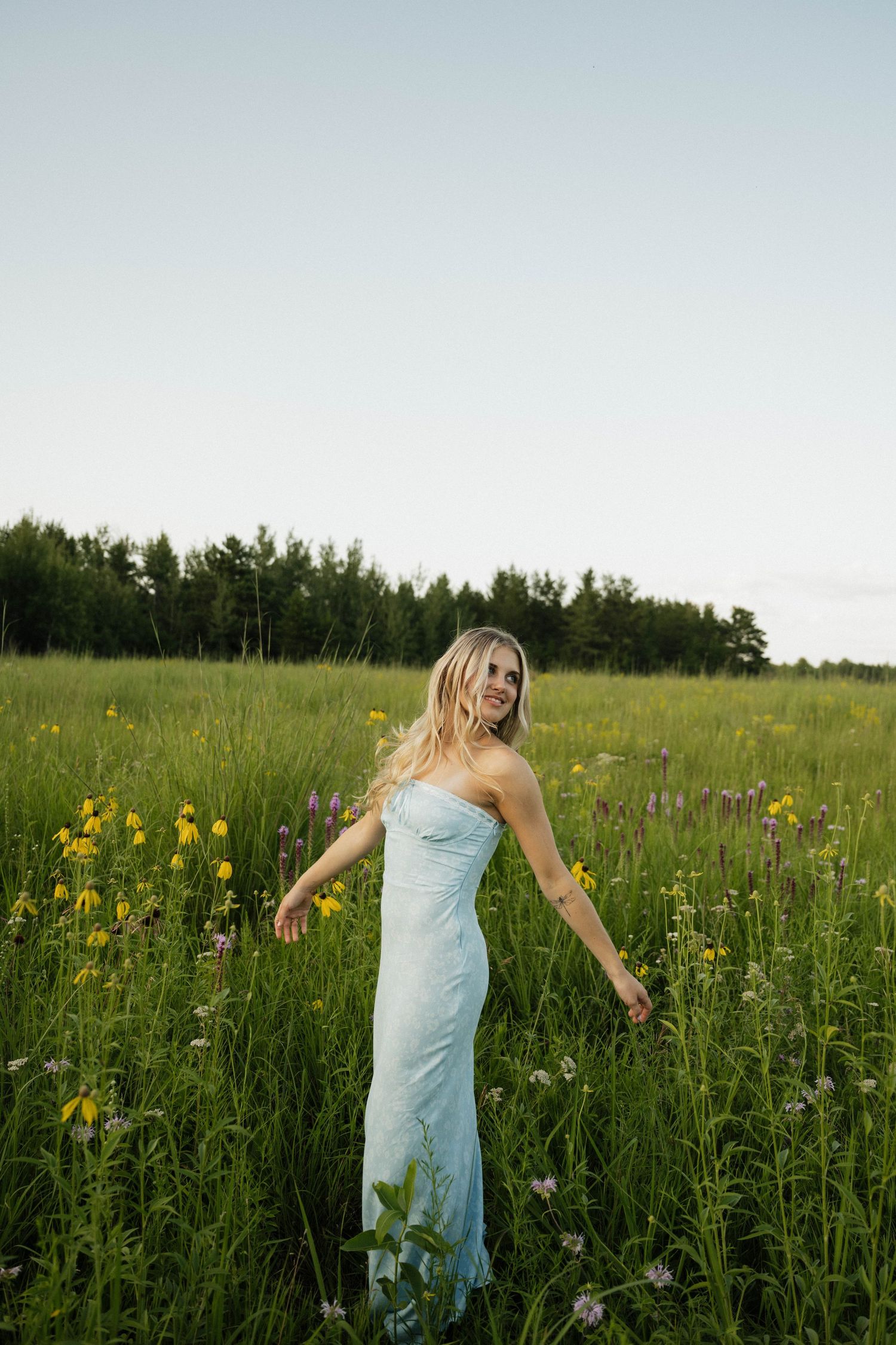 A smiling blonde woman in a light blue strapless gown walks through a lush wildflower meadow at dusk.