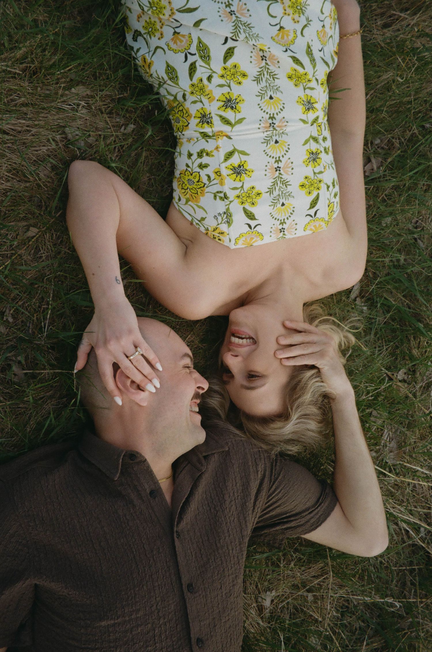Couple laughing and lying on grass, woman in floral dress and man in brown shirt, aerial view.