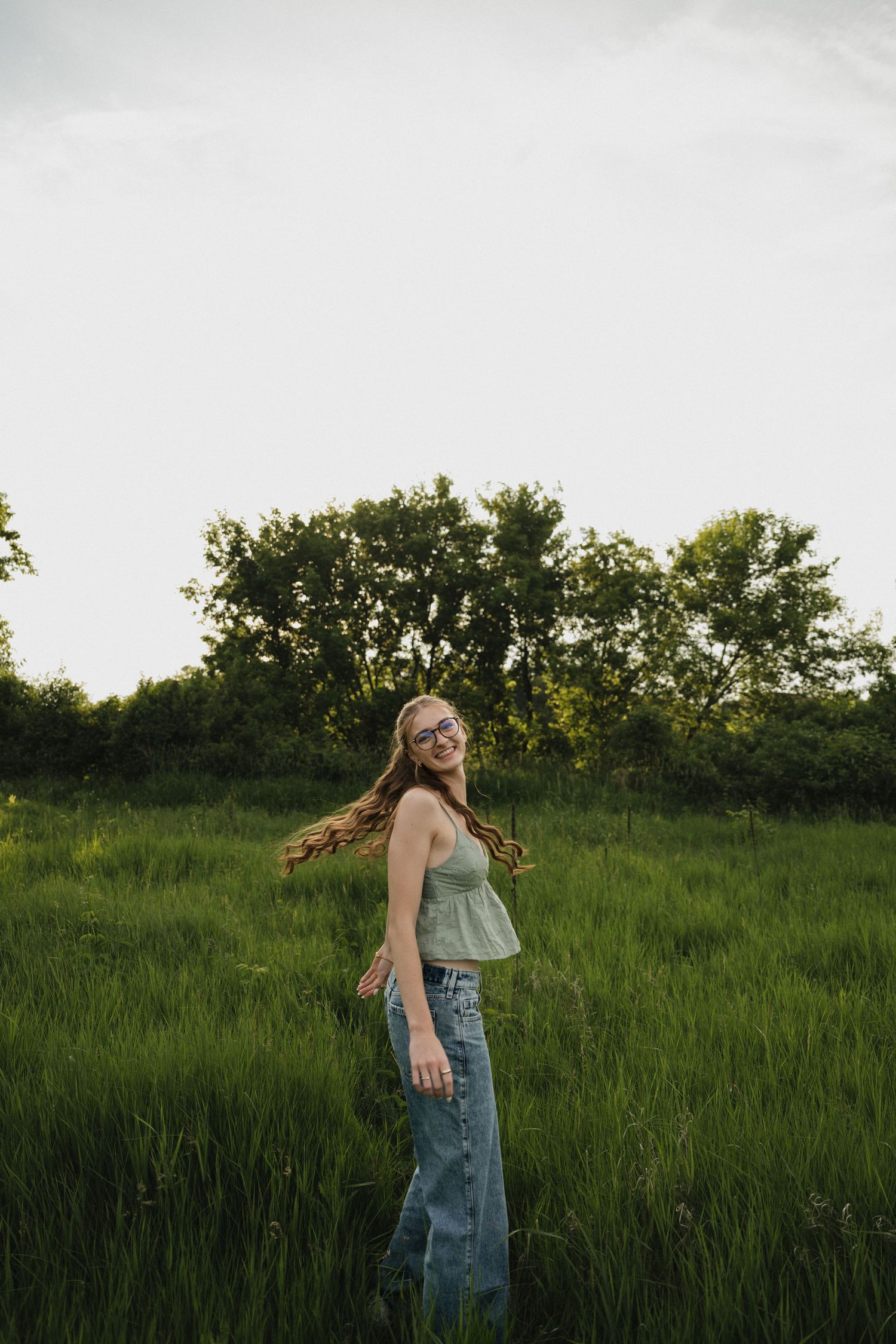 Young woman with long hair smiling in a green field, wearing a sage top and jeans, with trees in the background.