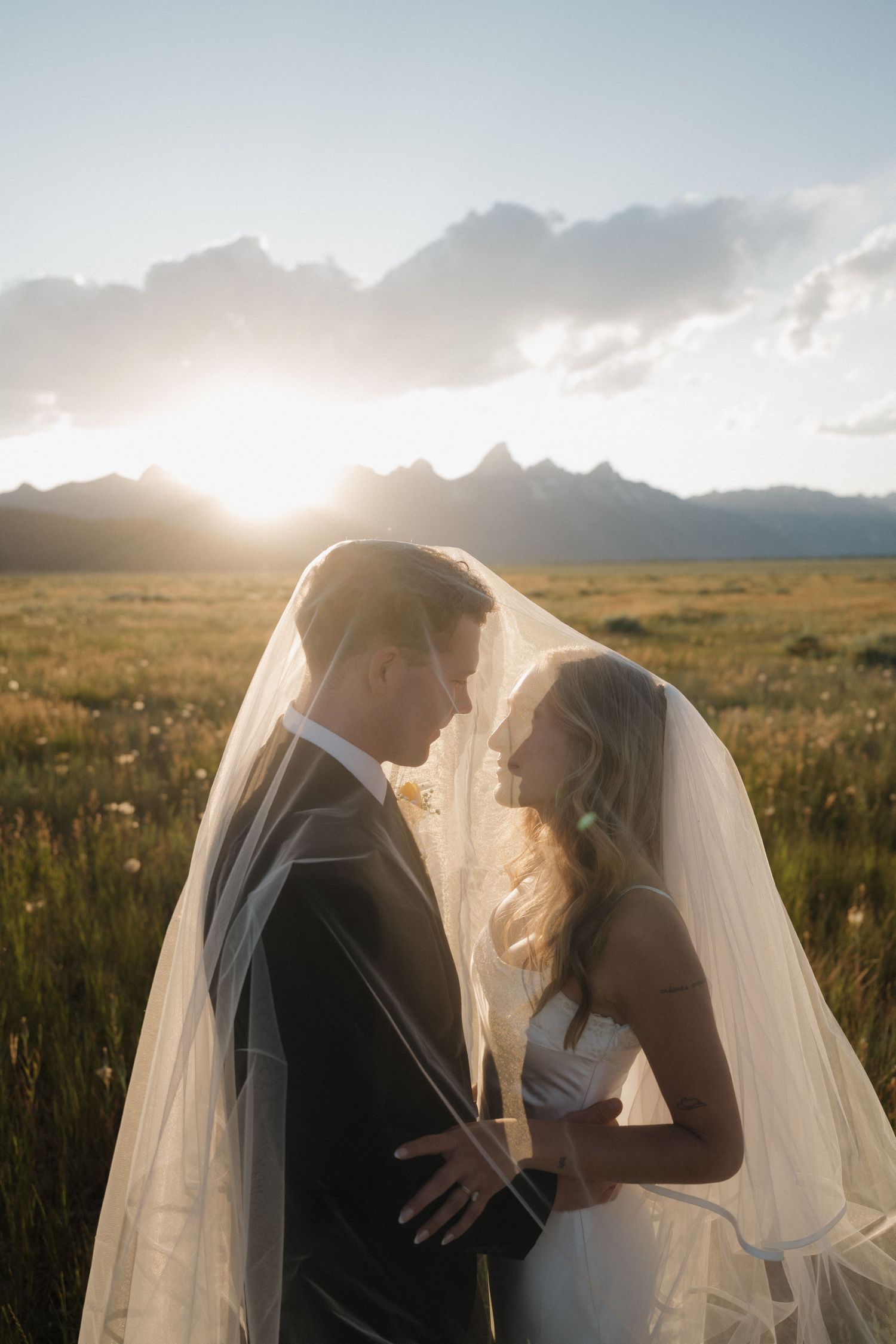 Bride and groom share romantic moment under veil in golden sunlit mountain field at sunset.