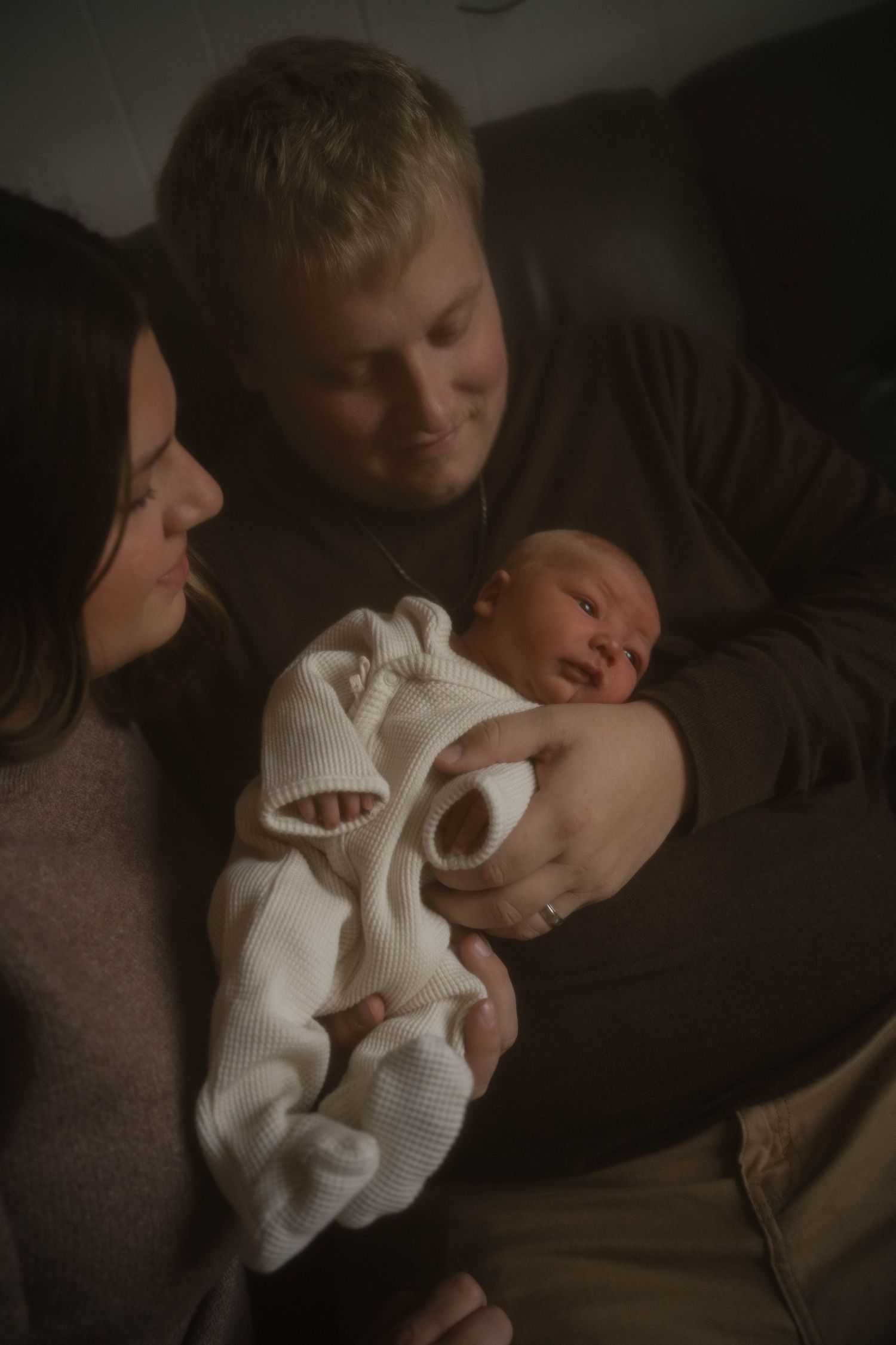 New parents tenderly holding their newborn baby dressed in white in a dimly lit room.