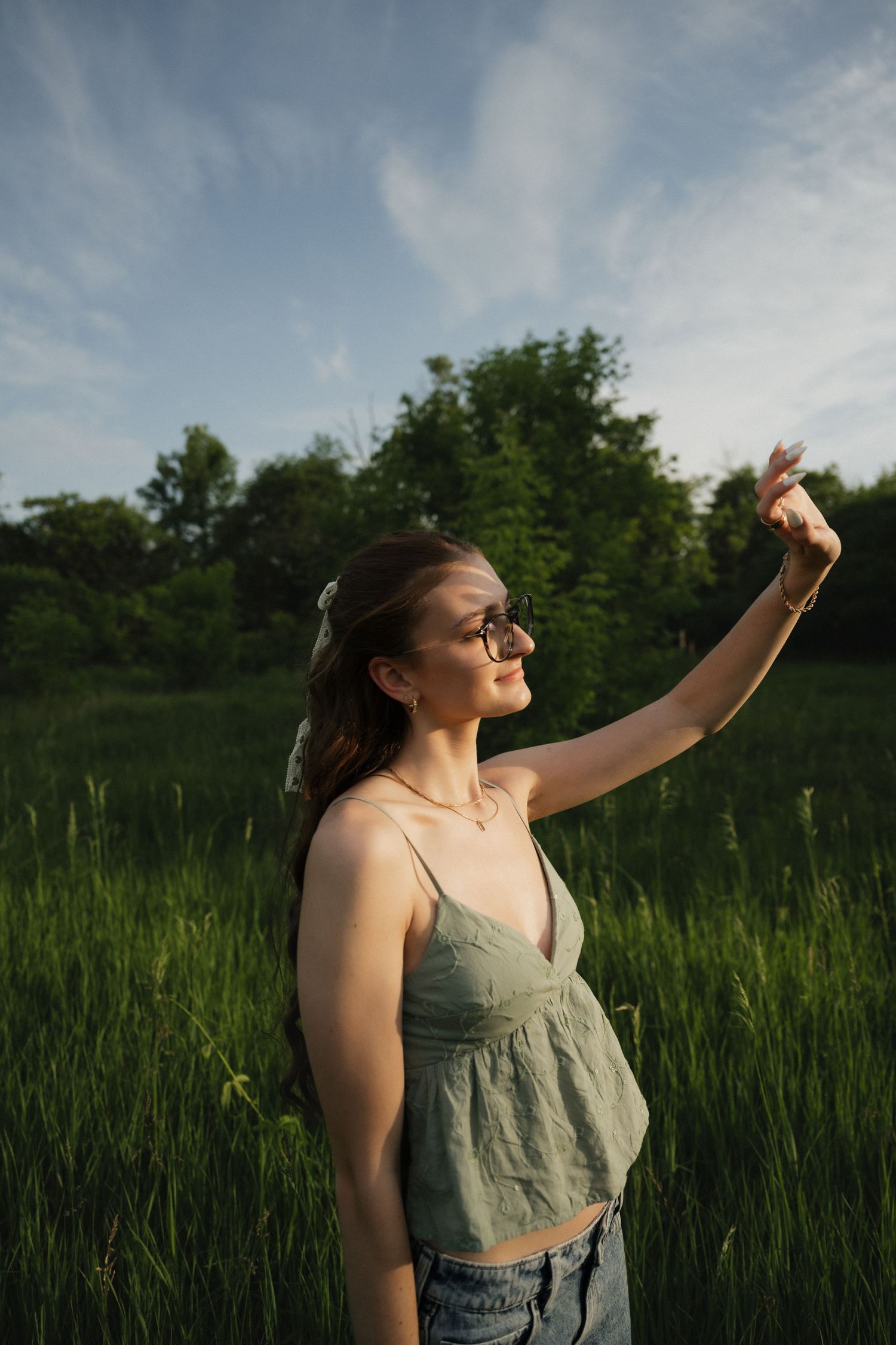 Smiling woman in sage green crop top and sunglasses raising hand in sunlit green meadow.