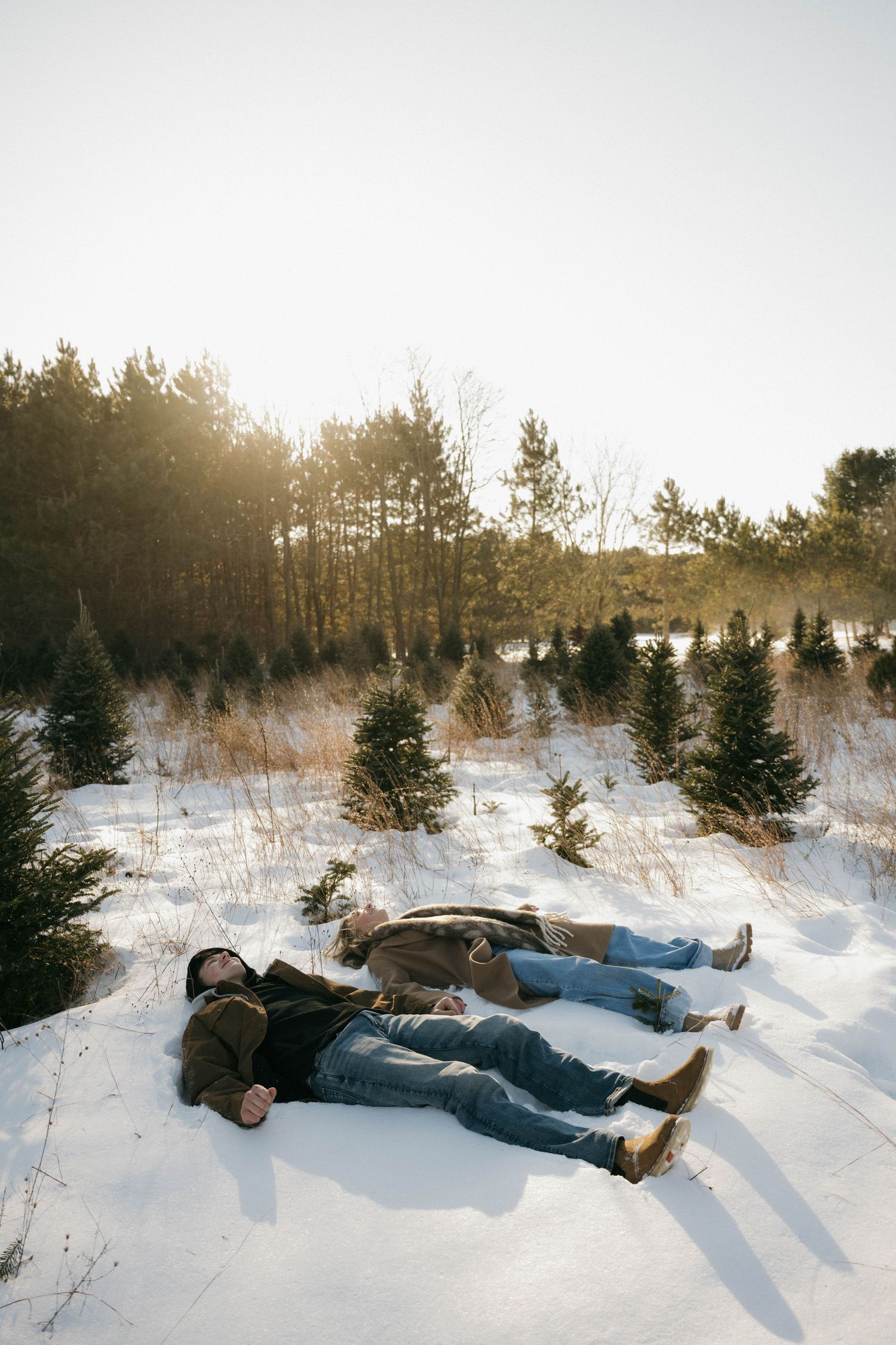 Couple lying in the snow making snow angels at a winter Christmas tree farm.