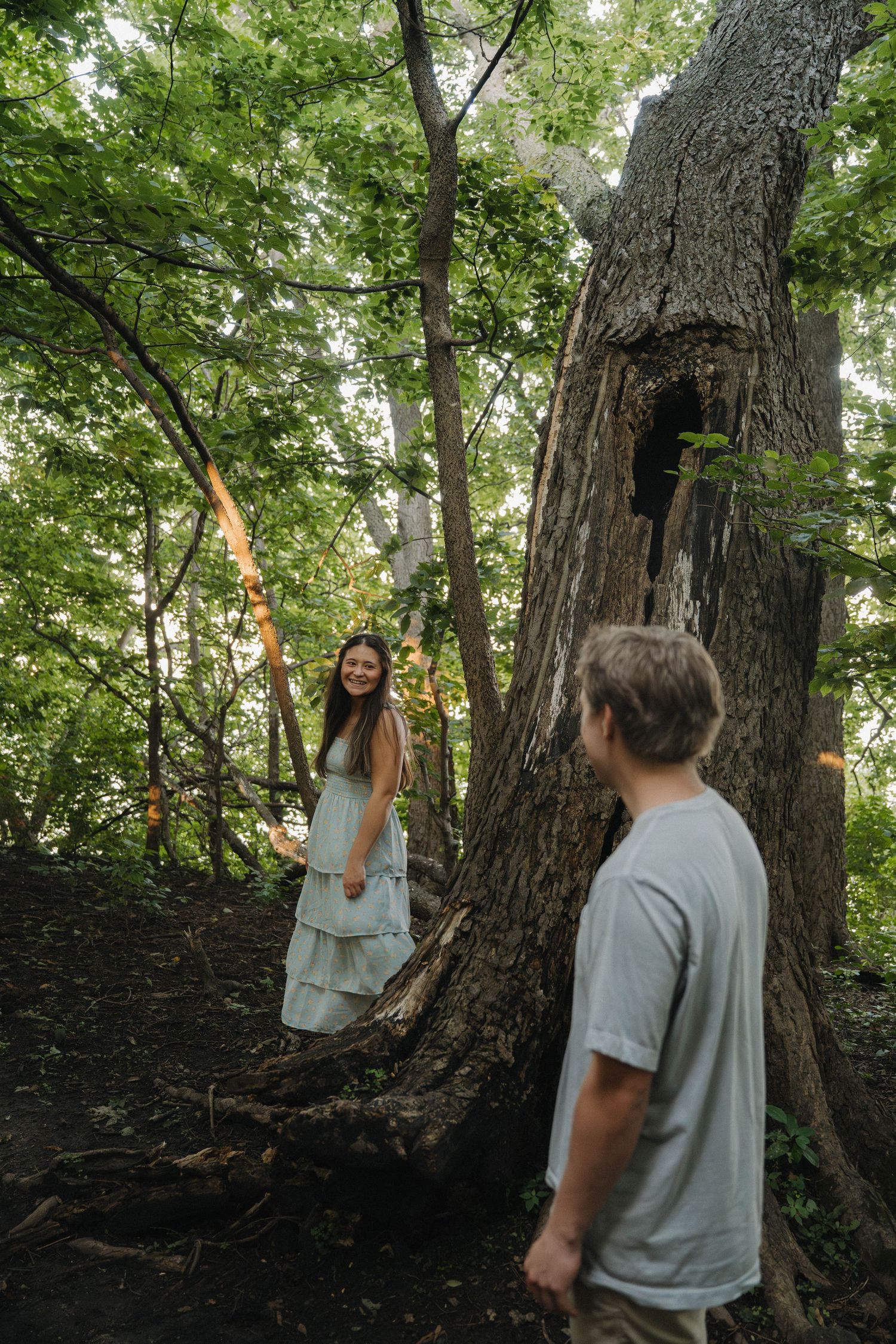 Young woman in tiered blue dress smiling by large tree in forest, man in gray shirt in foreground.
