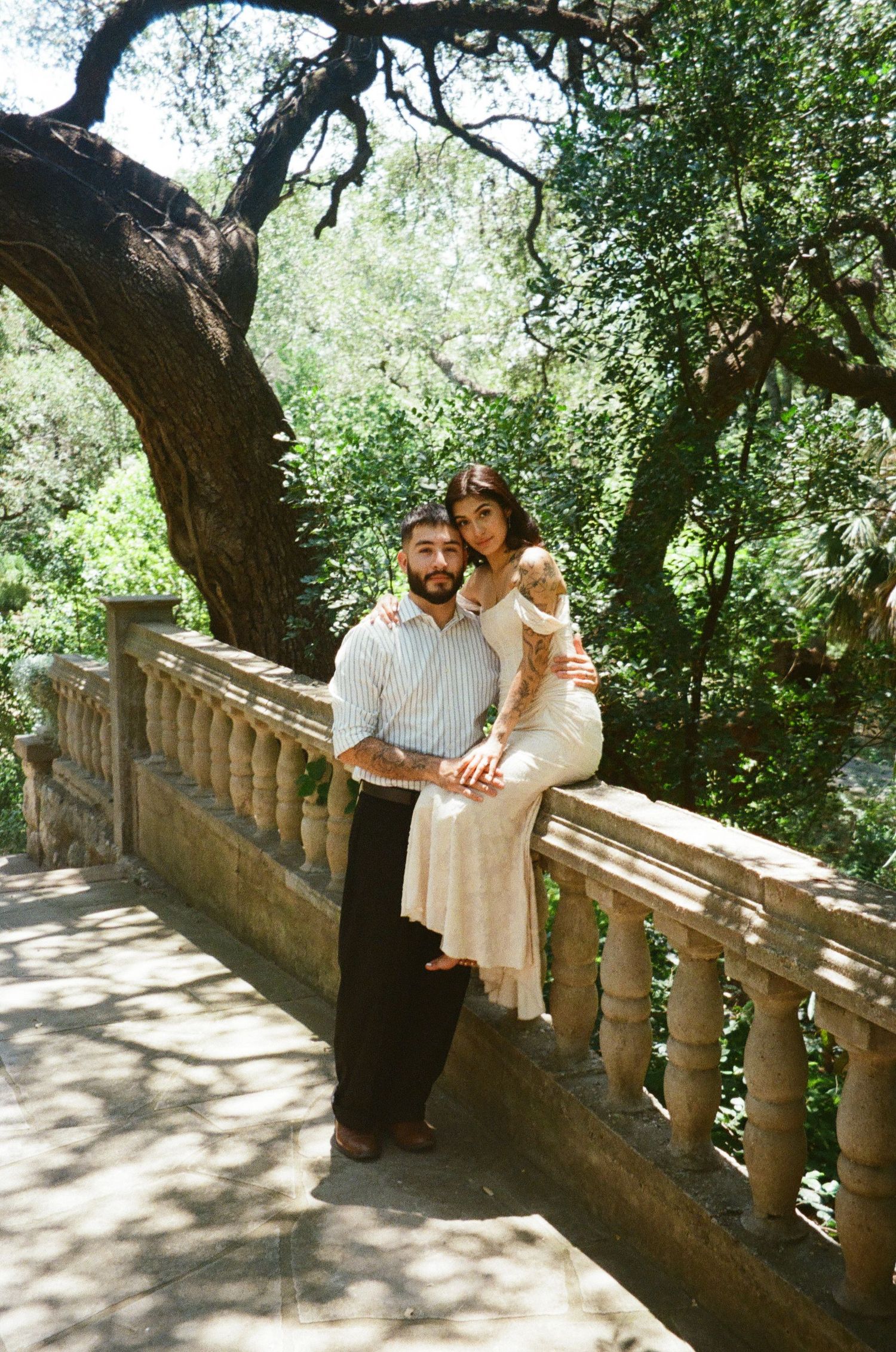 Tattooed couple posing together on stone balcony railing surrounded by lush green trees.