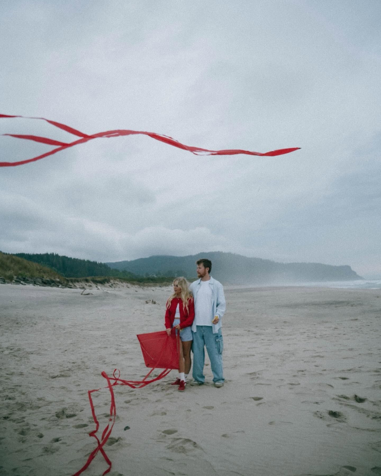 Couple standing on a sandy beach flying a red kite on a cloudy day with misty mountains in the background.