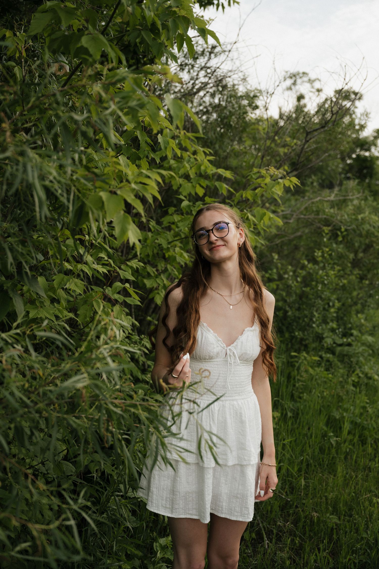 Young woman in white sundress and glasses smiling among lush green trees and foliage outdoors.