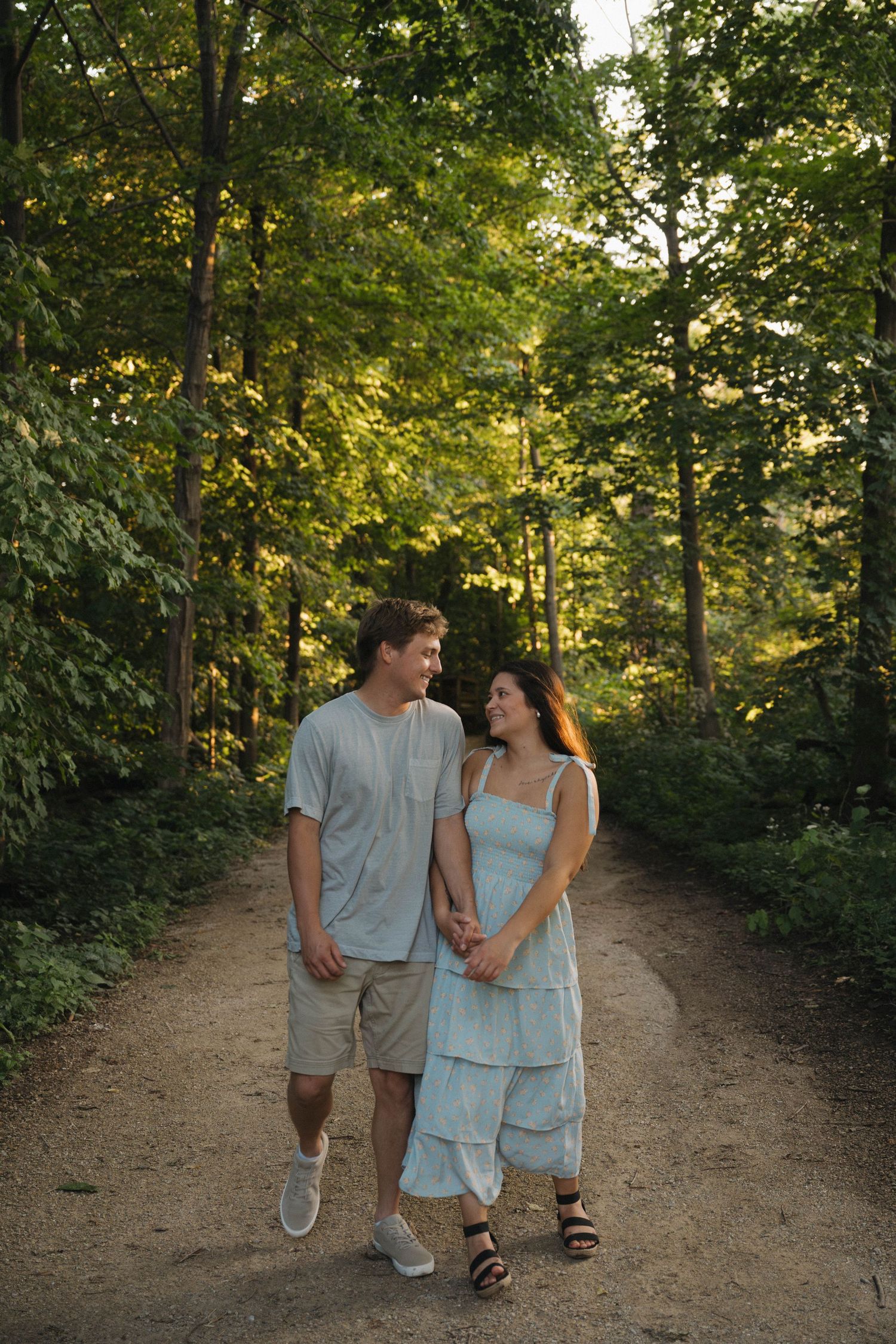 A couple holds hands and smiles at each other while walking on a forest path surrounded by lush green trees.