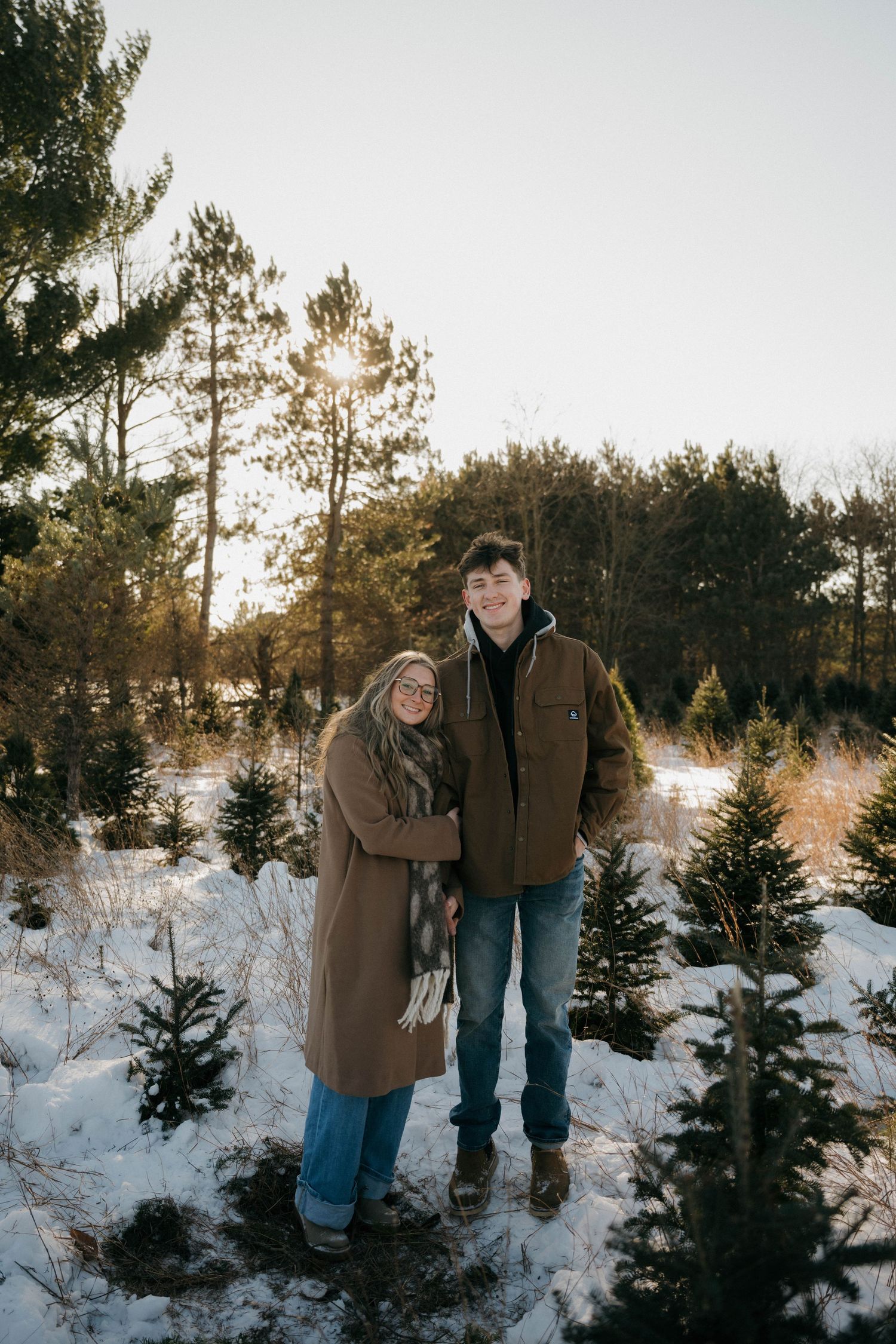 A smiling couple poses together at a snowy Christmas tree farm during golden hour in winter coats.