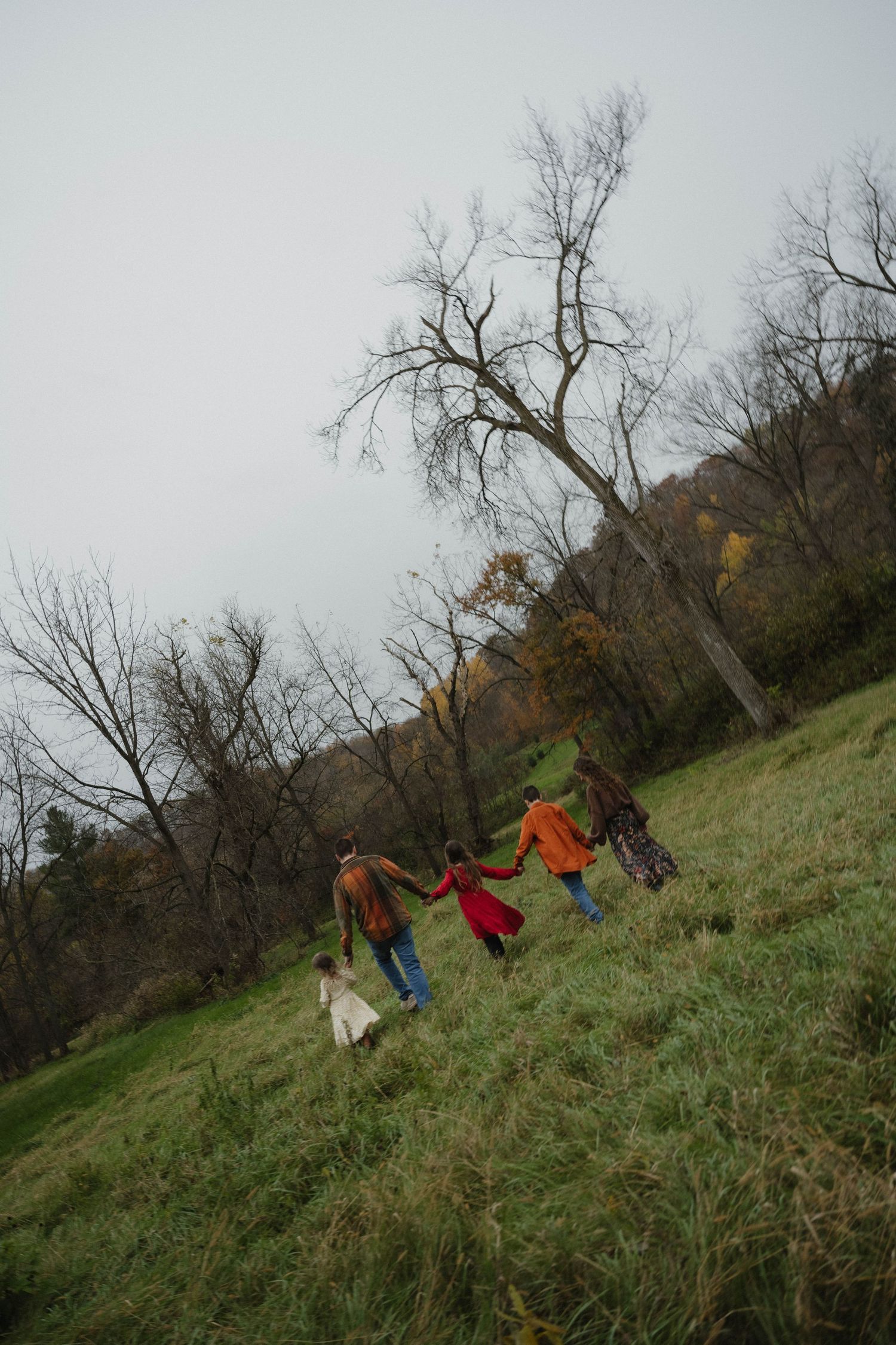 A family of five holding hands walking up a green hillside with bare autumn trees in background.