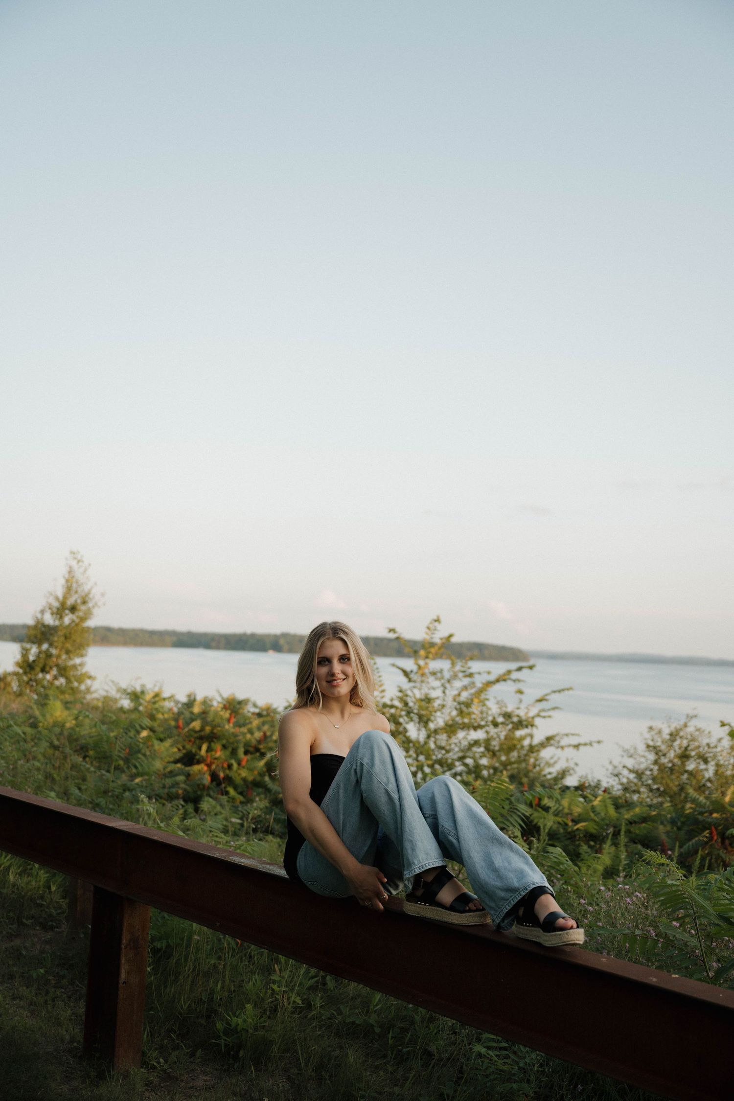A smiling blonde woman sits on a wooden rail overlooking a serene lake at dusk.